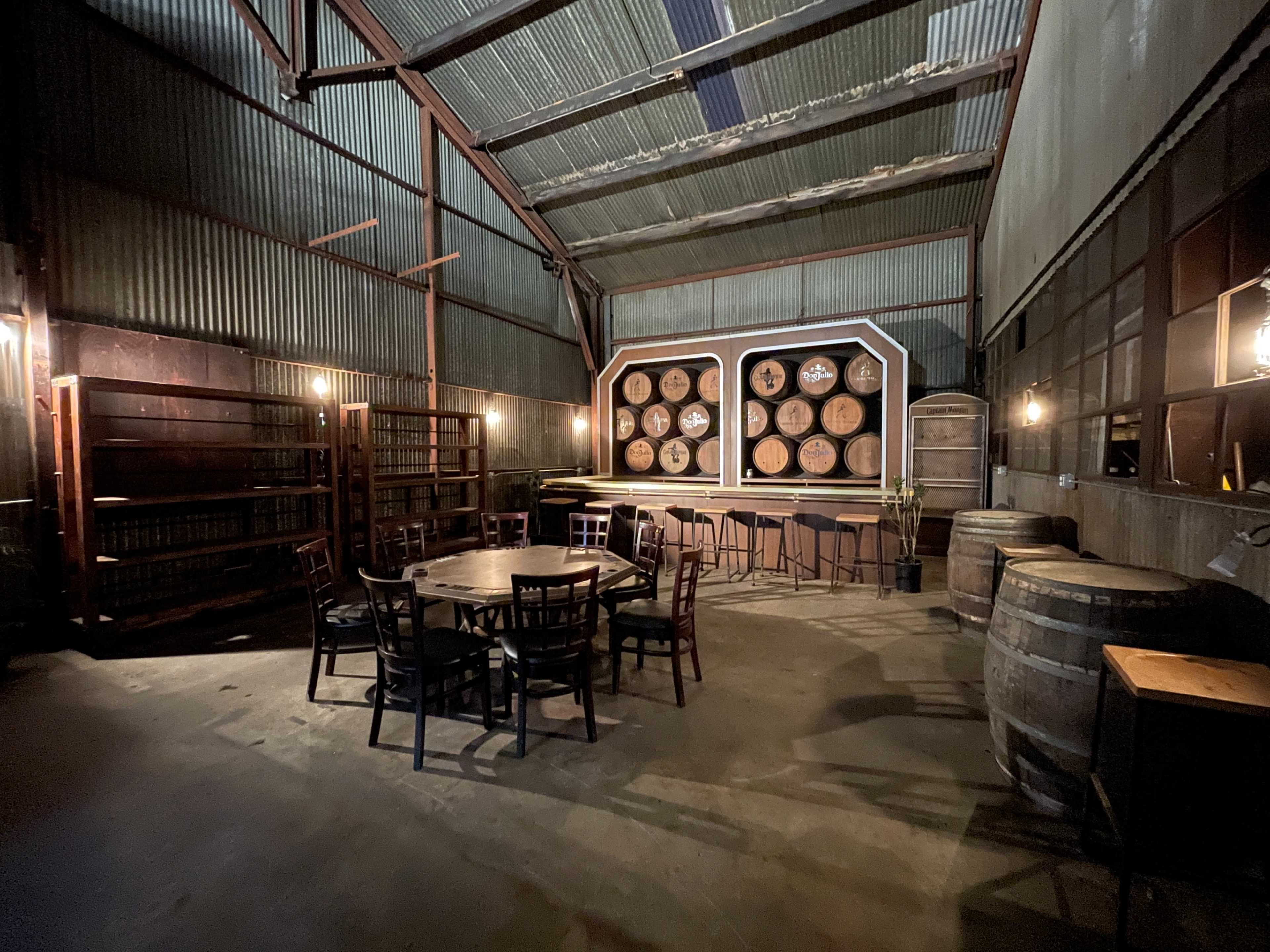 The interior of a rustic warehouse features a large display of barrels on the back wall, surrounded by empty tables and chairs.