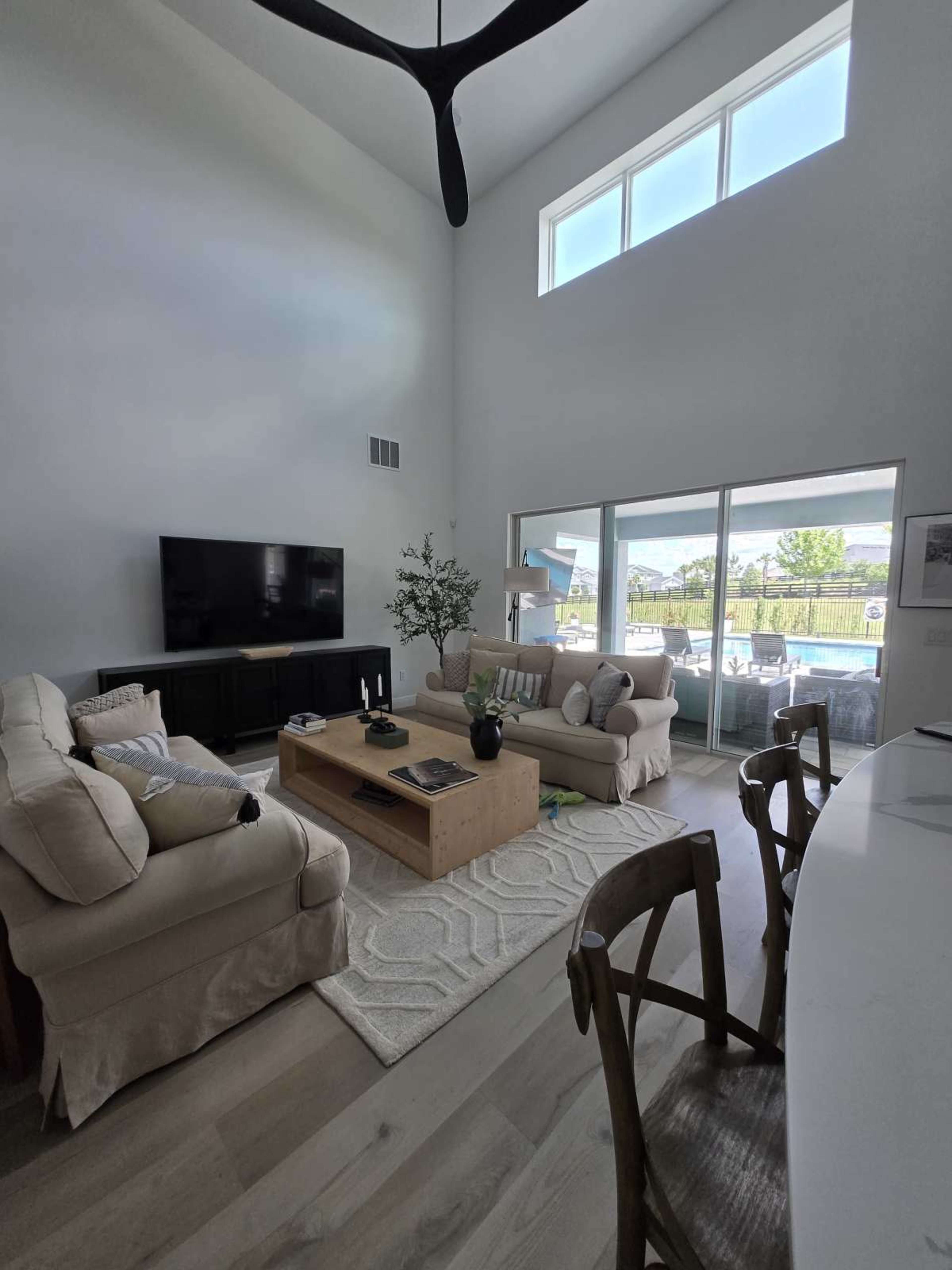 The image shows a bright living room with a high ceiling, featuring a television, a light-colored sofa, a coffee table, and large windows that overlook a patio area.