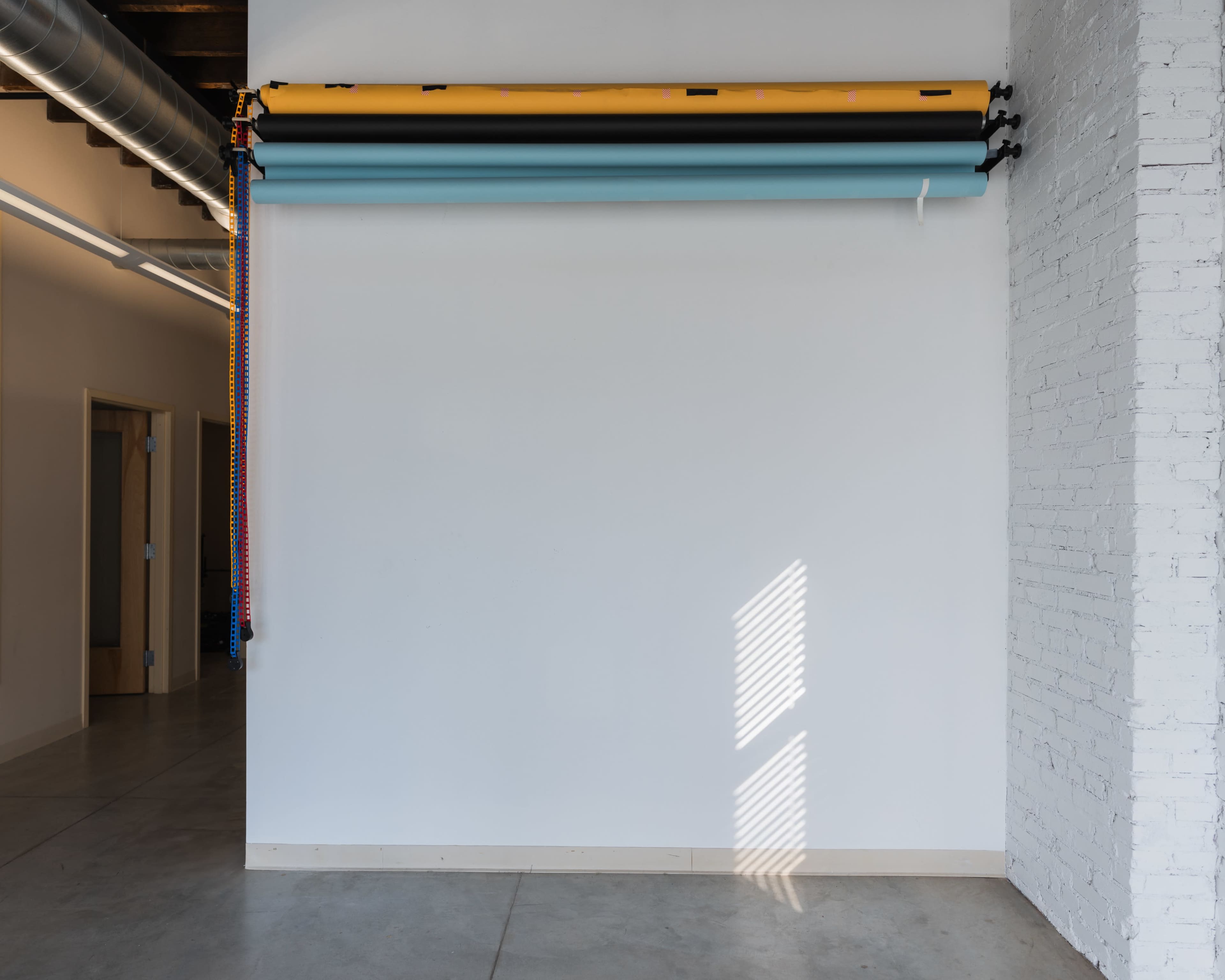 A white wall in a spacious room displays rolled-up backgrounds in yellow and blue, with sunlight casting shadows on the floor.