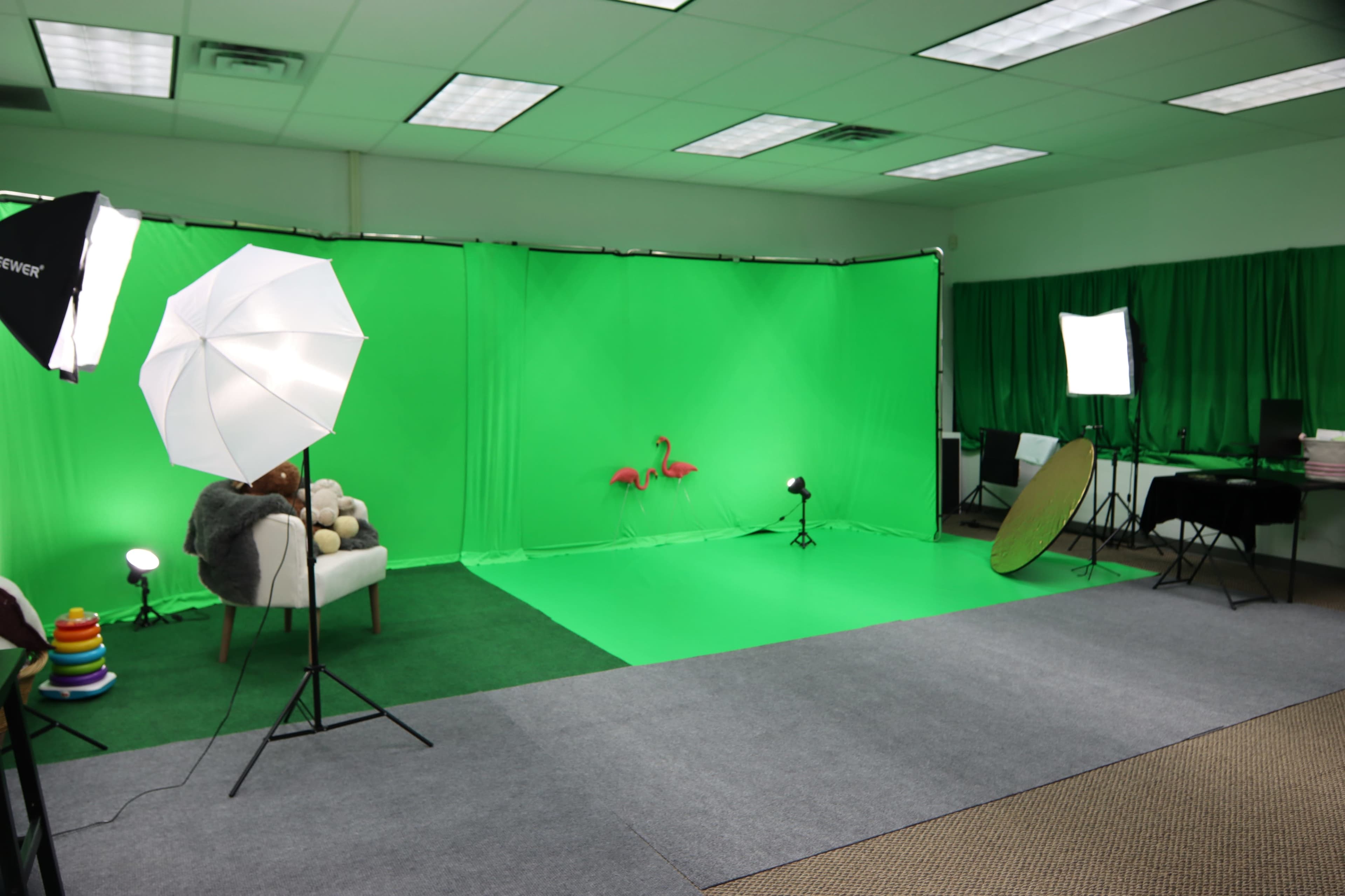 A green screen studio set up with photography lights, a vanity mirror, and a seating area featuring a plush chair and some decorative items.