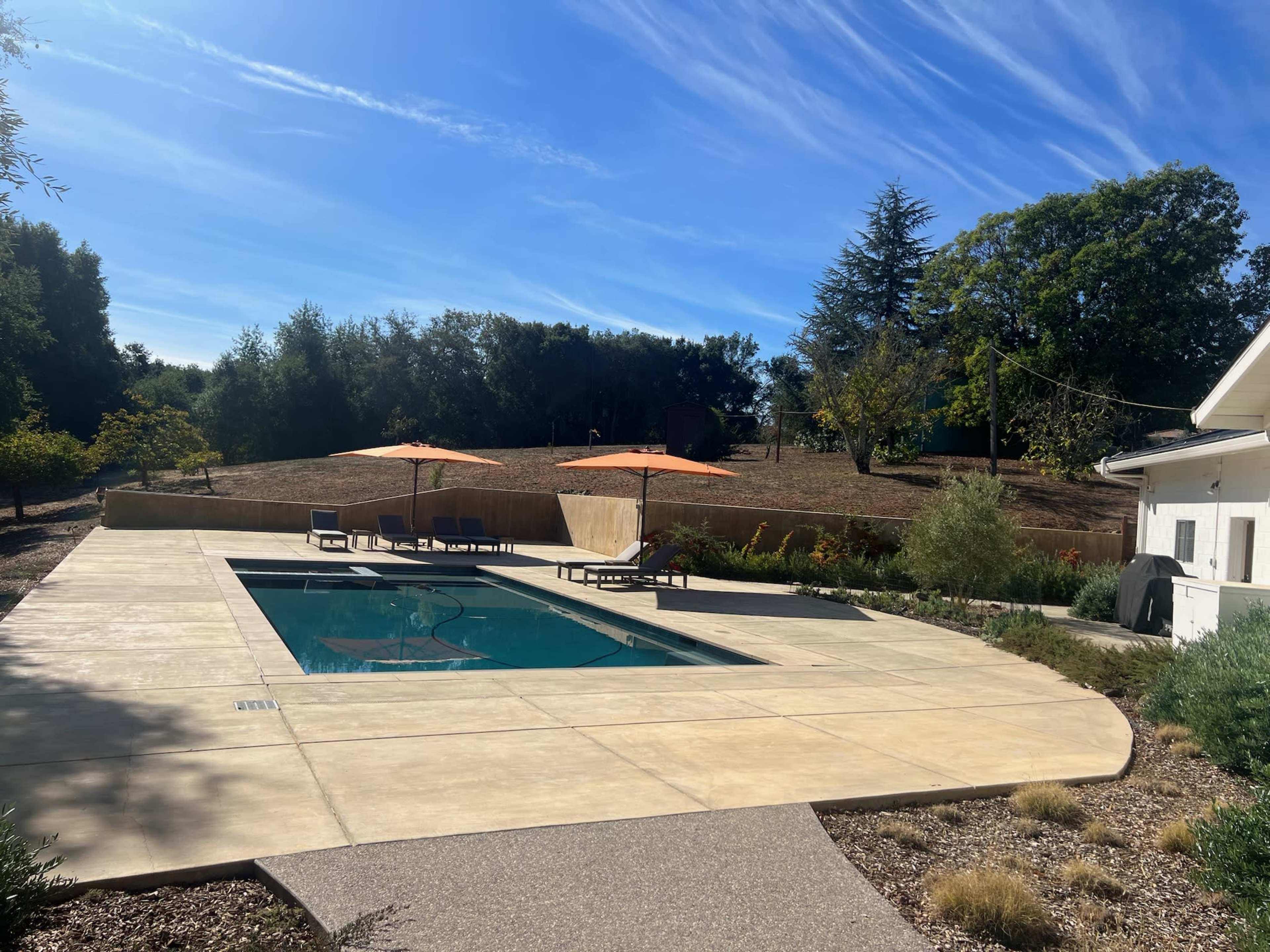 The image shows a rectangular swimming pool surrounded by a spacious concrete deck and lounge chairs, with orange umbrellas, set against a backdrop of trees and a clear blue sky.