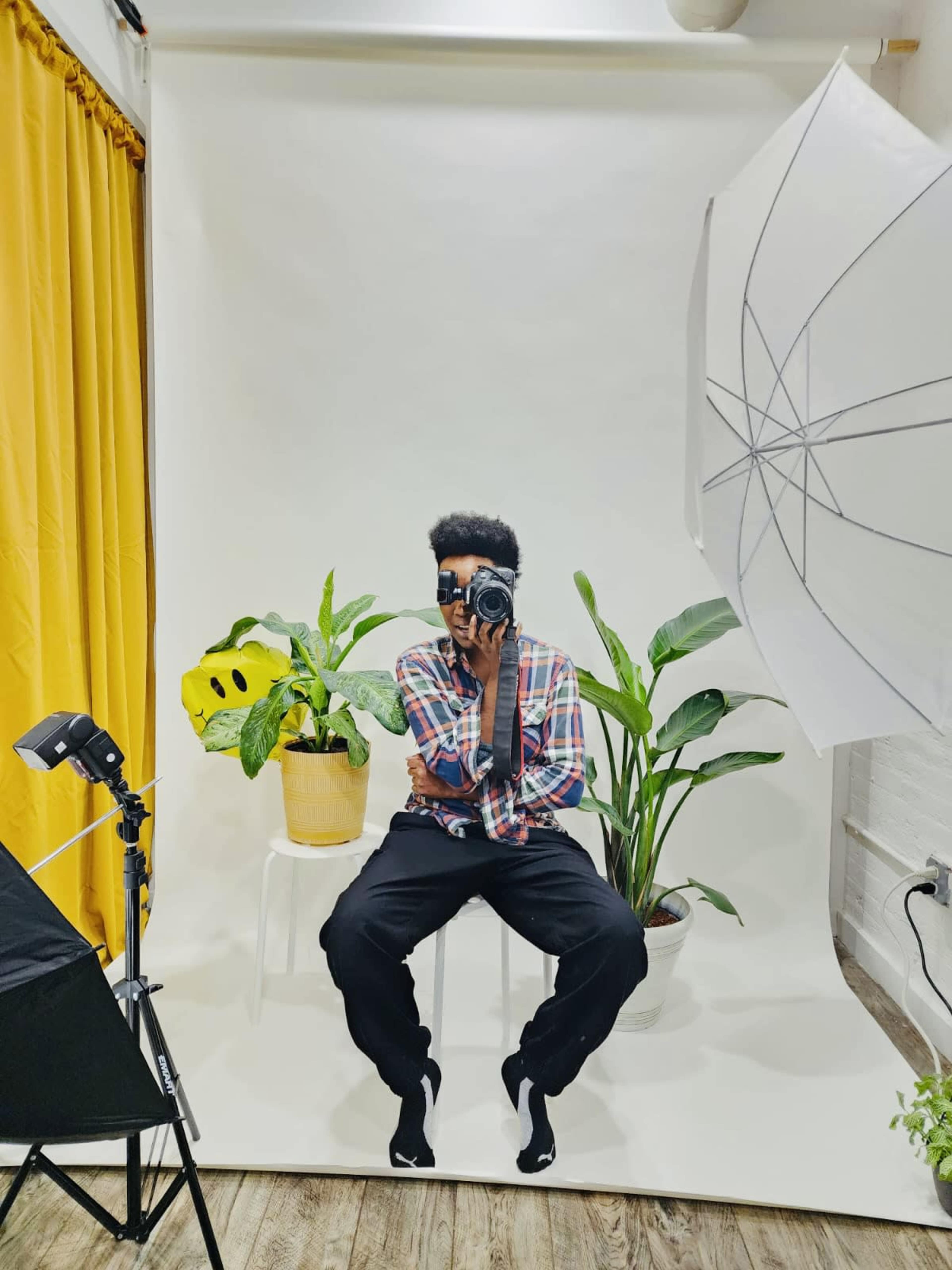A person is sitting on a white stool in front of a light backdrop, holding a camera, surrounded by potted plants and studio lighting equipment.