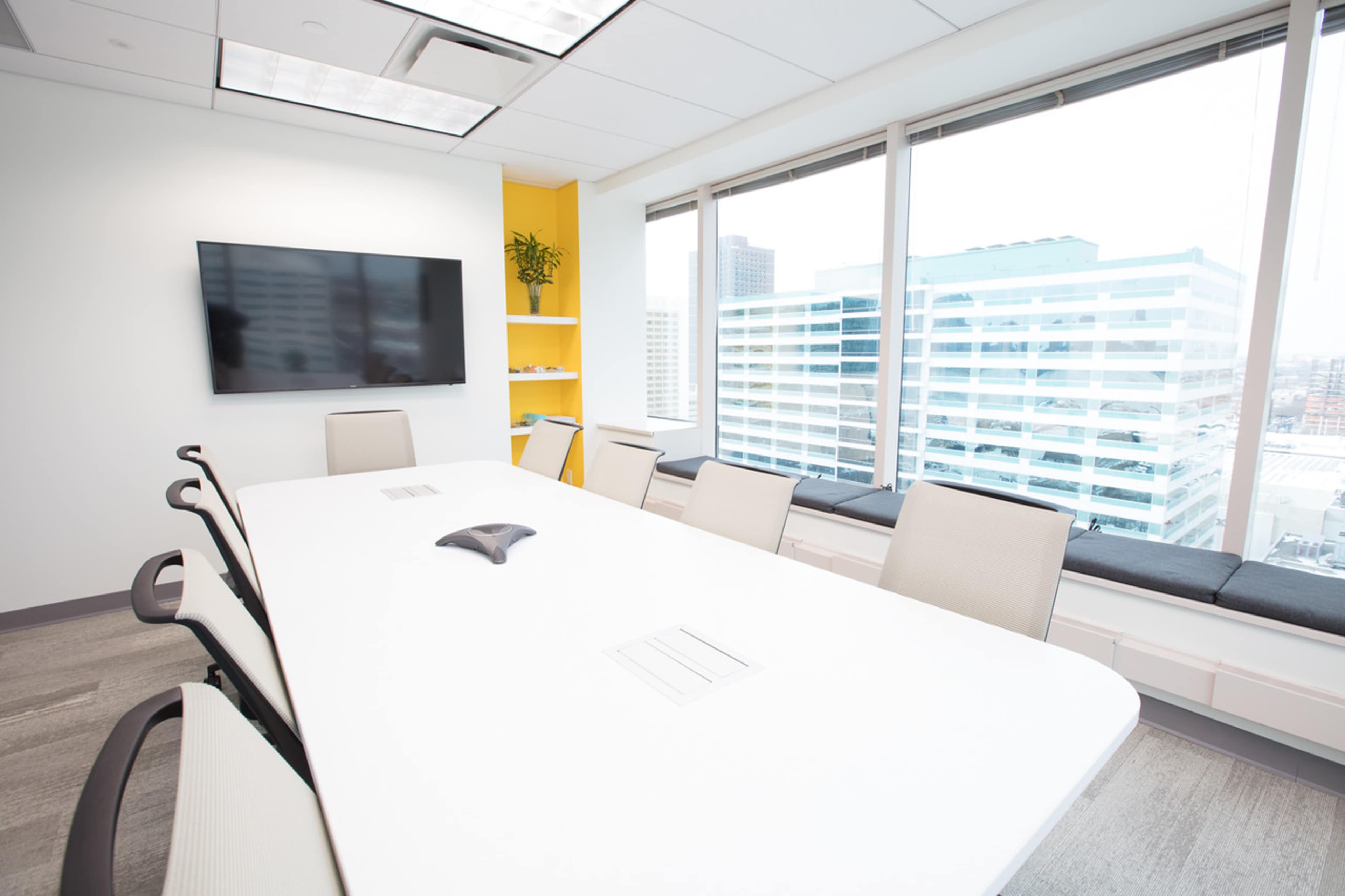 A modern conference room features a long white table surrounded by light-colored chairs, with a large screen mounted on the wall and large windows overlooking a cityscape.