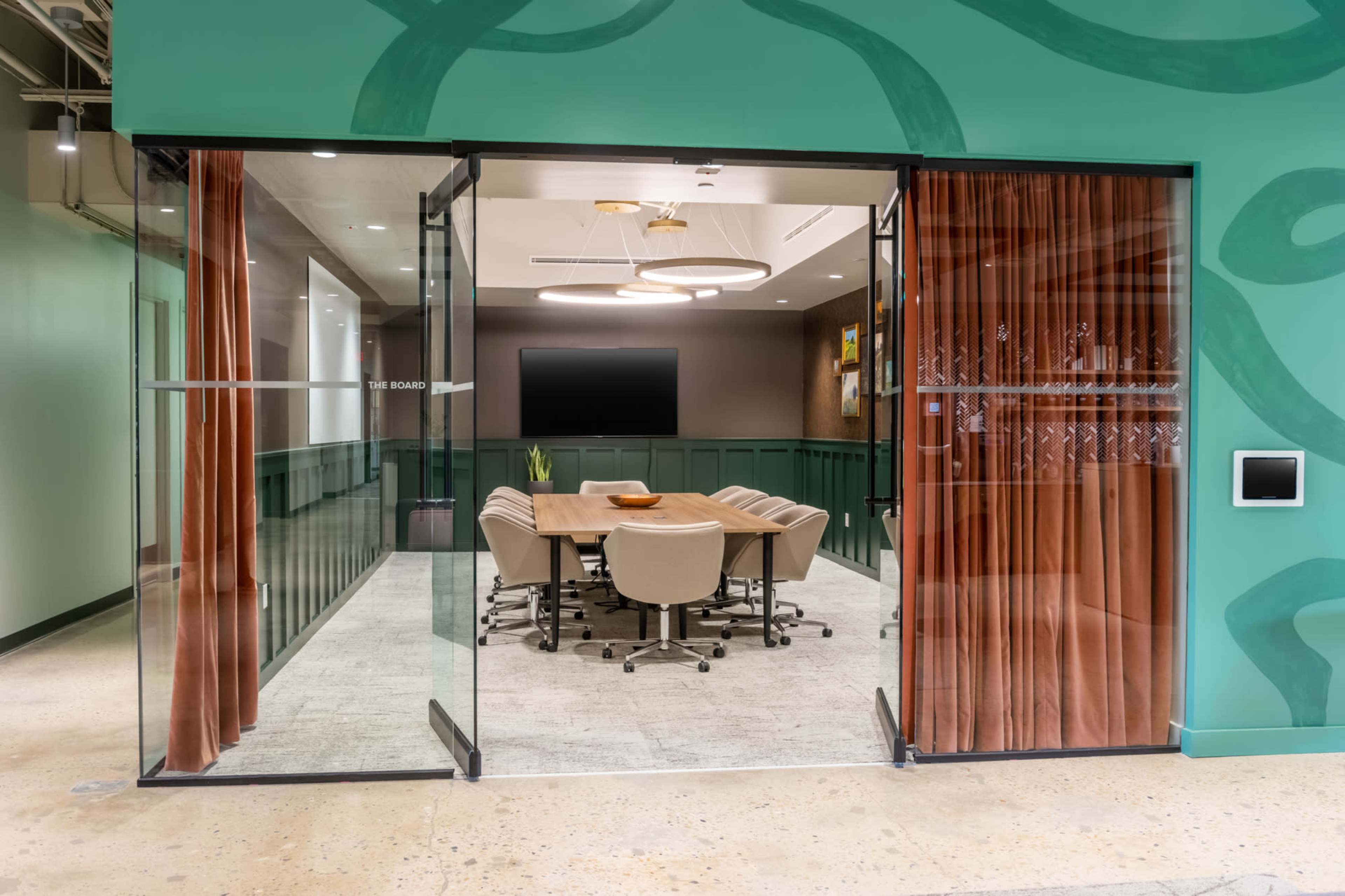 A conference room with a large table and beige chairs is visible through glass doors, featuring green walls and curtain drapes.