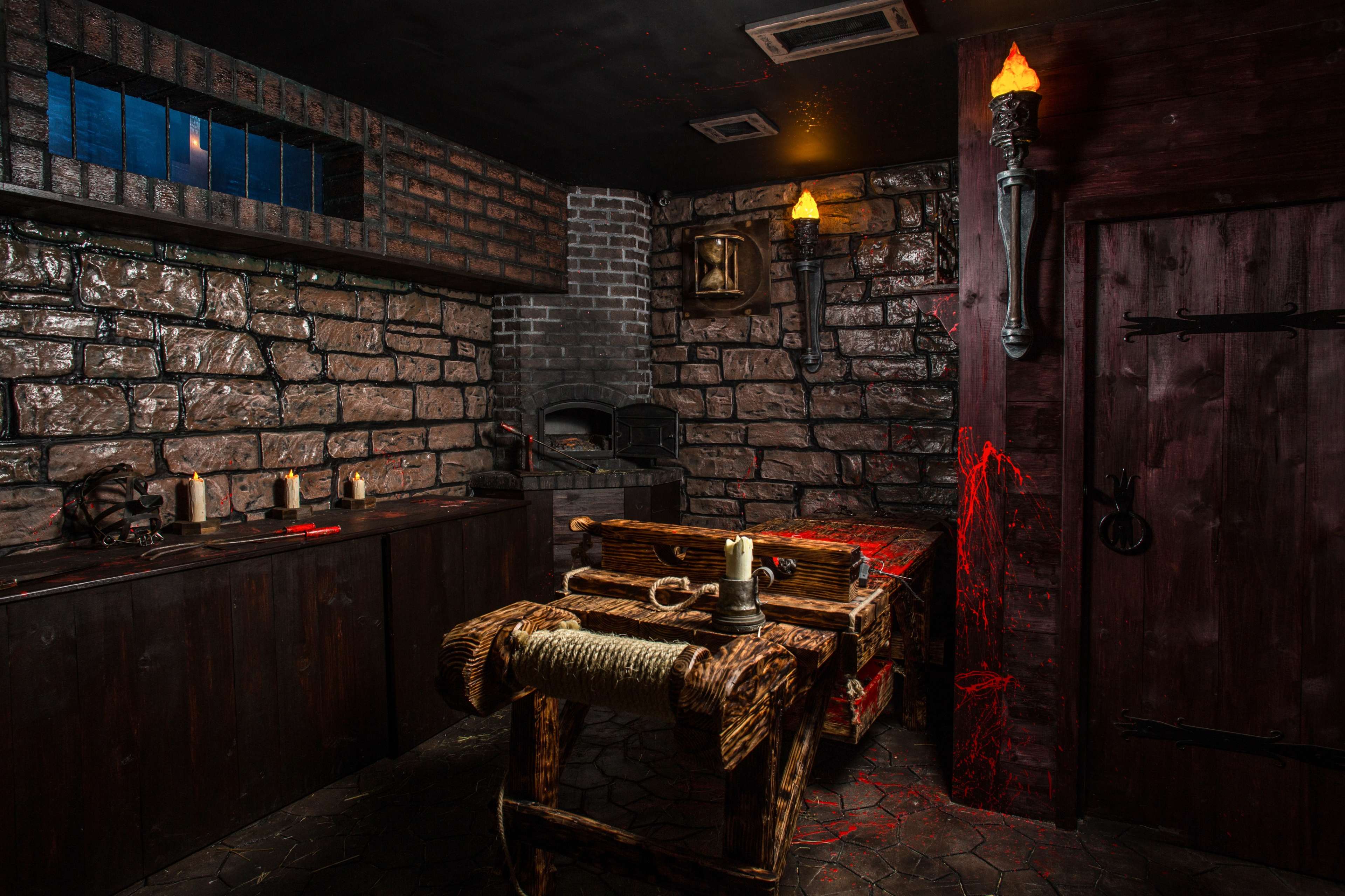 A dimly lit dungeon room with stone walls, a wooden torture table in the center, and a brick oven in the background.