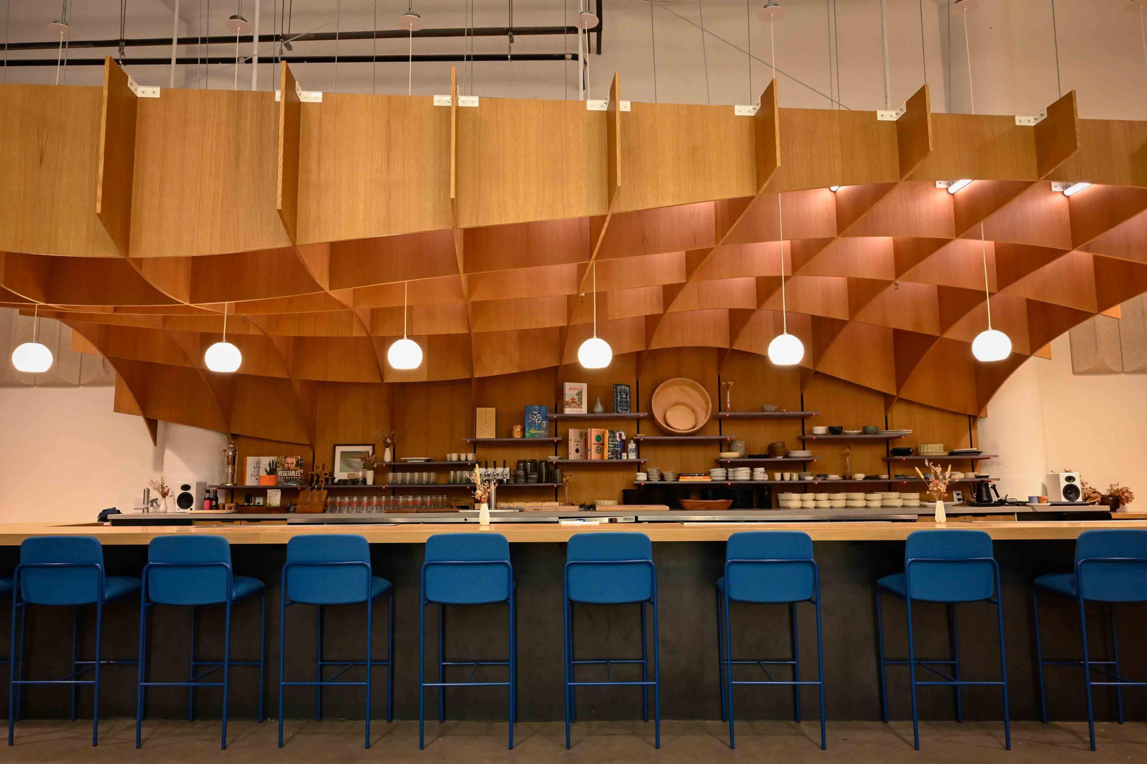 A modern café features a wooden ceiling design above a long counter with blue stools and decorative items on shelves.