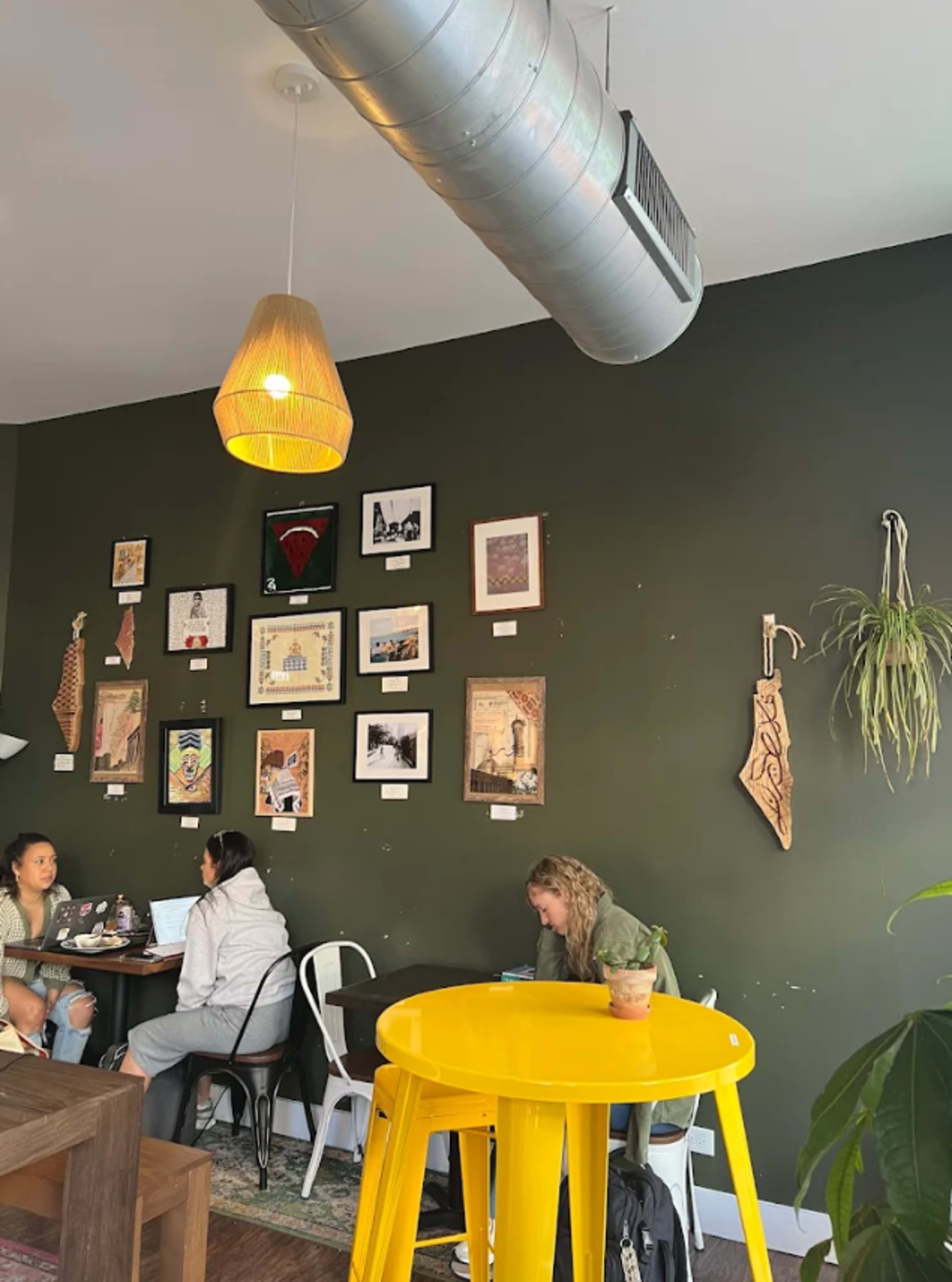 The image shows a cozy café interior featuring a green wall adorned with various framed artworks, while two groups of people, one sitting at a table and another at a yellow table, engage in conversation and work on laptops.