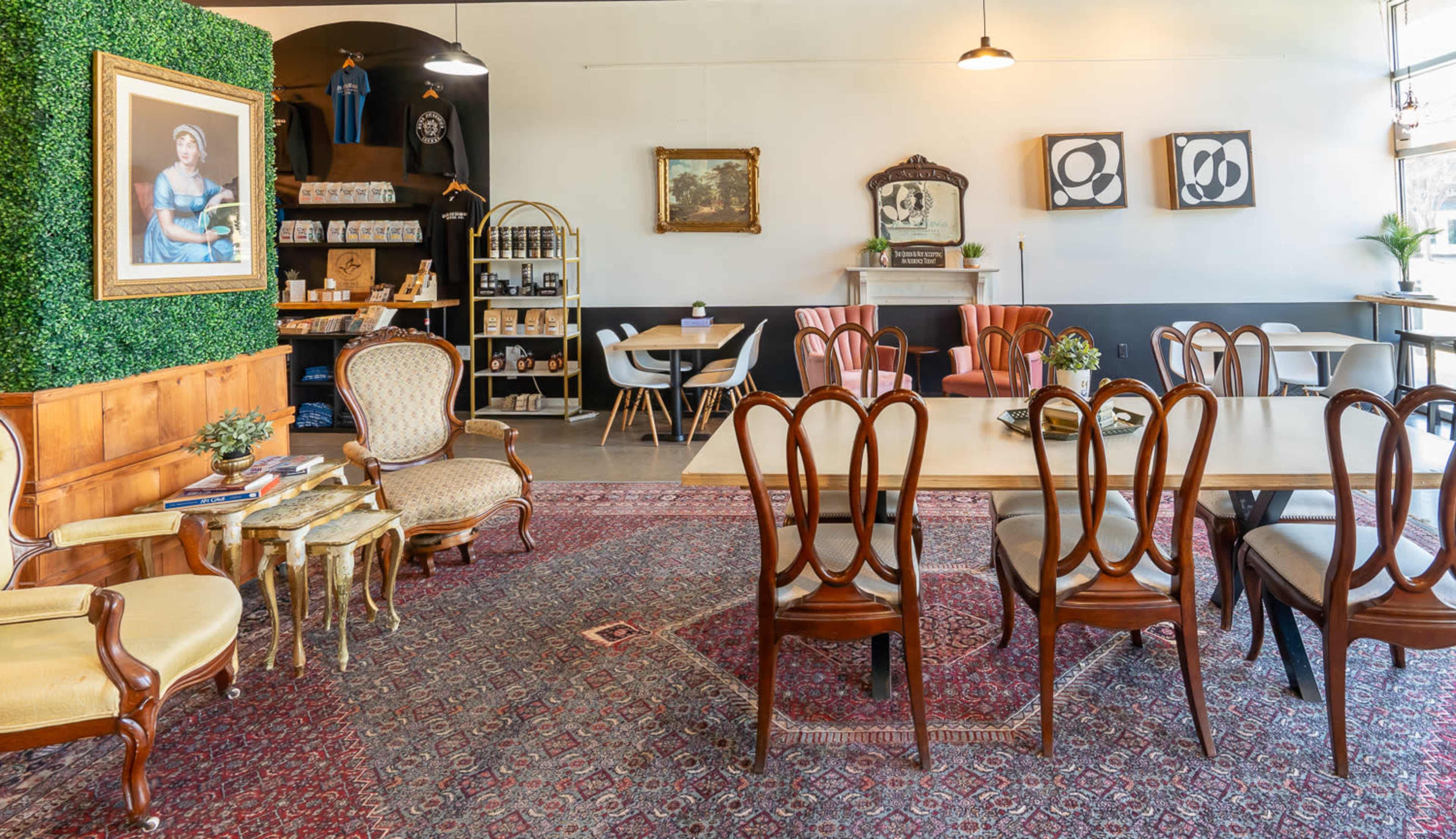A cozy cafe interior featuring wooden furniture, a patterned rug, a decorative wall with plants, and framed artwork.