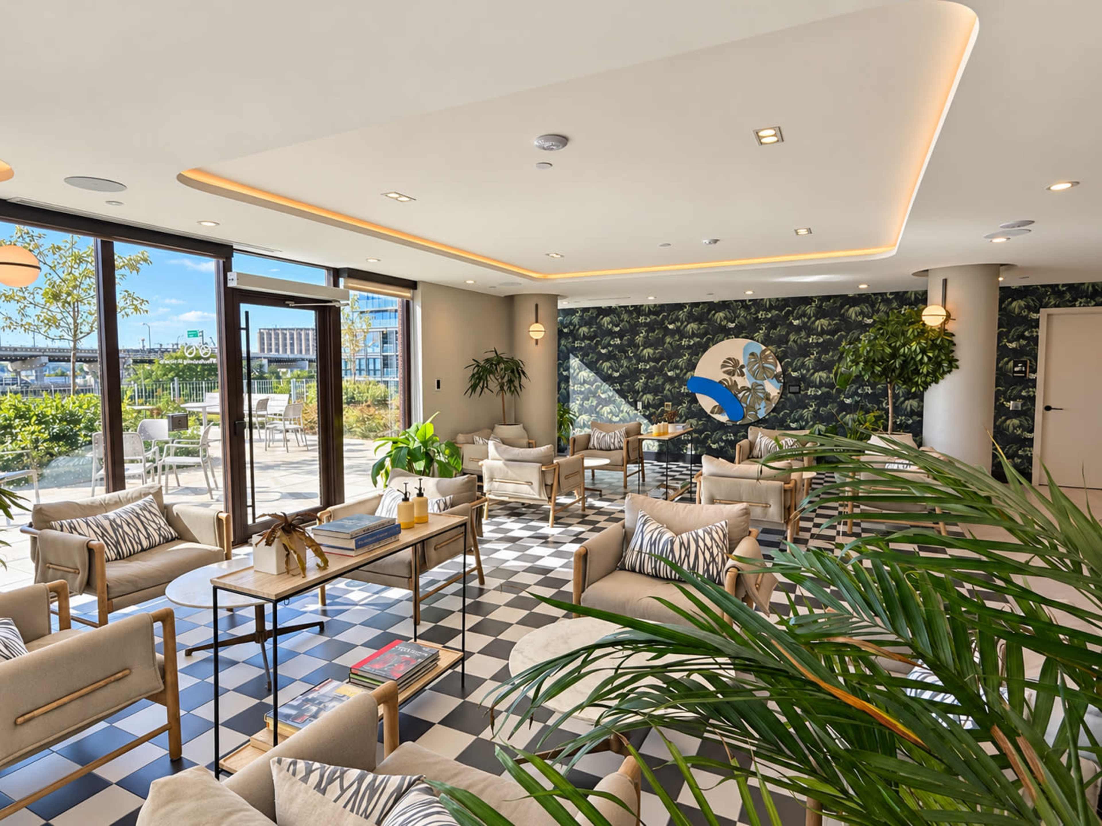 The image shows a modern lounge area with seating arranged around coffee tables, adorned with plants and large windows that allow natural light to fill the space.