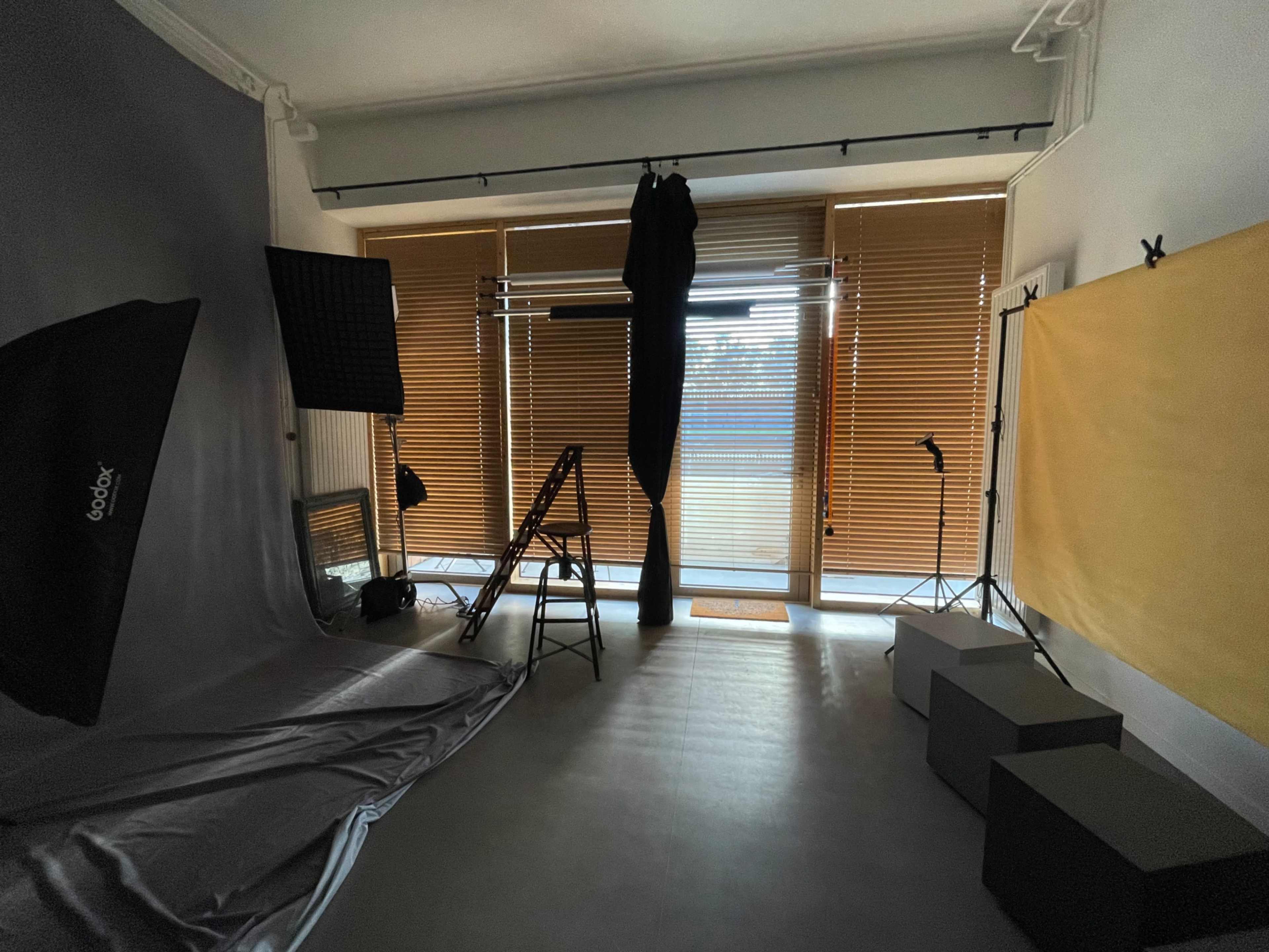 A photography studio with equipment, including softboxes, a backdrop, and a setup for lighting, along with large windows covered by blinds.