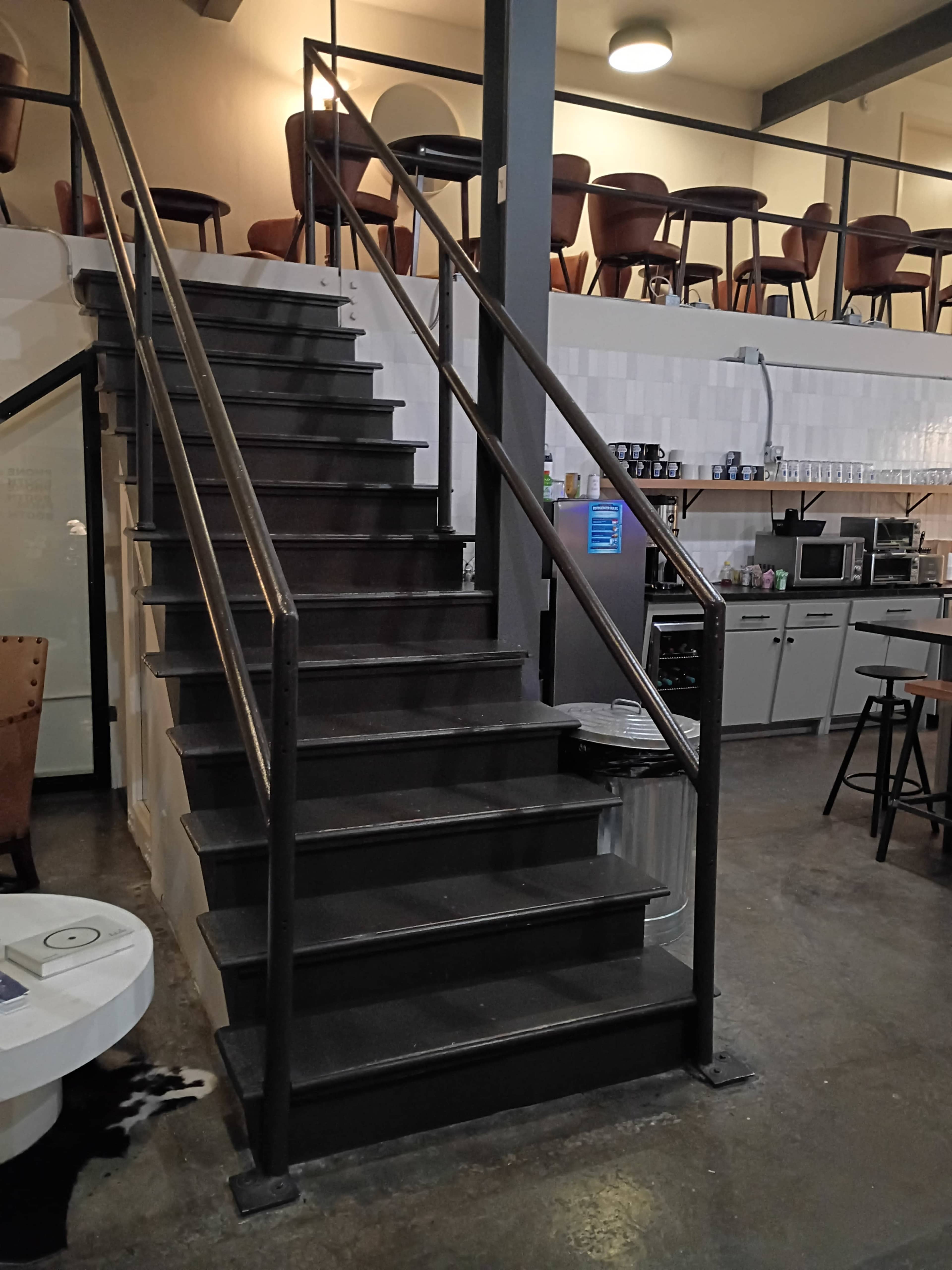 The image shows a set of metallic stairs leading to an upper level in a cafe, with chairs visible along the balcony and various kitchen appliances in the background.