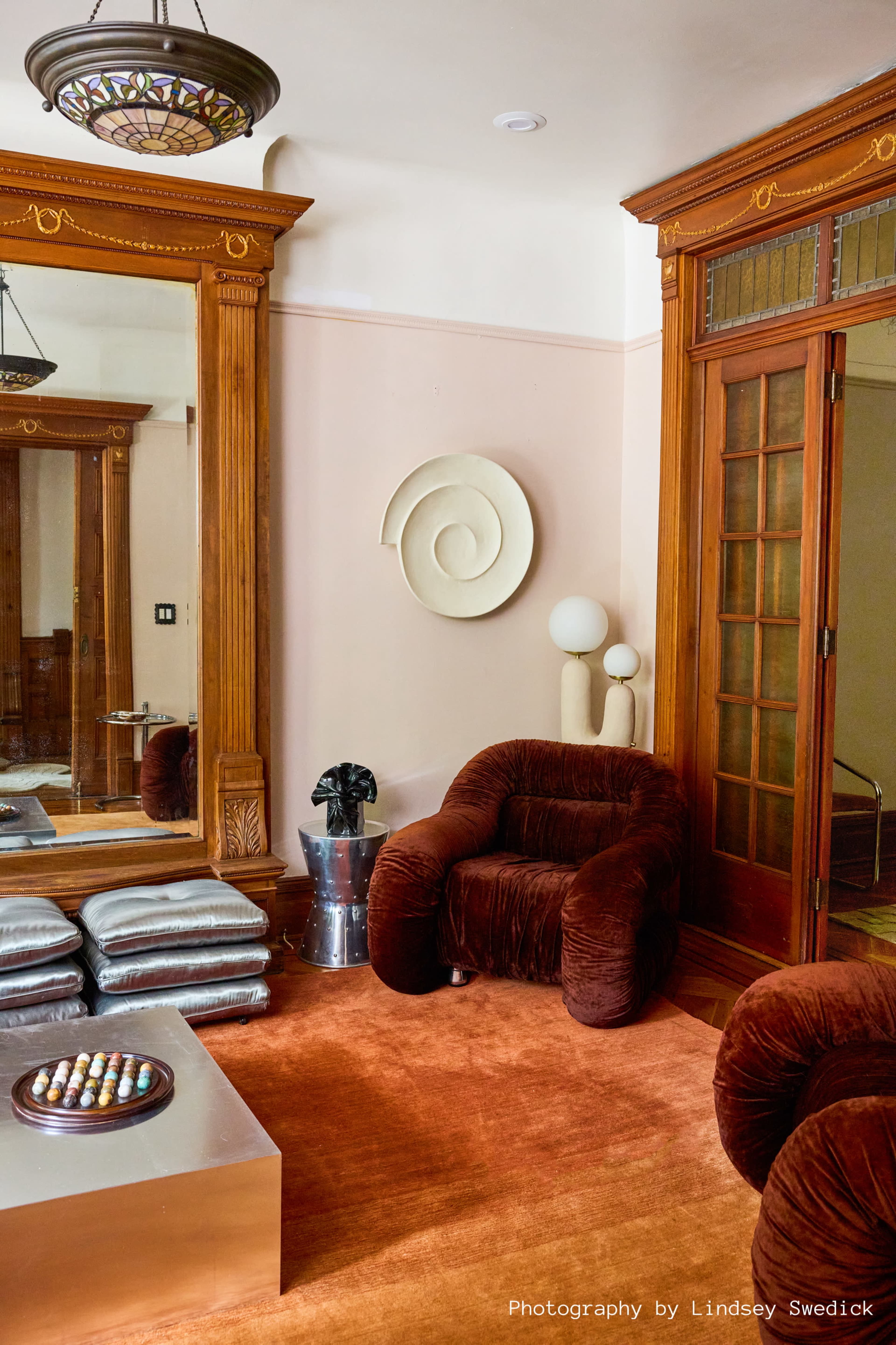The image shows a living room featuring a large mirror, plush seating, a circular wall art piece, and a metallic coffee table on a textured rug.