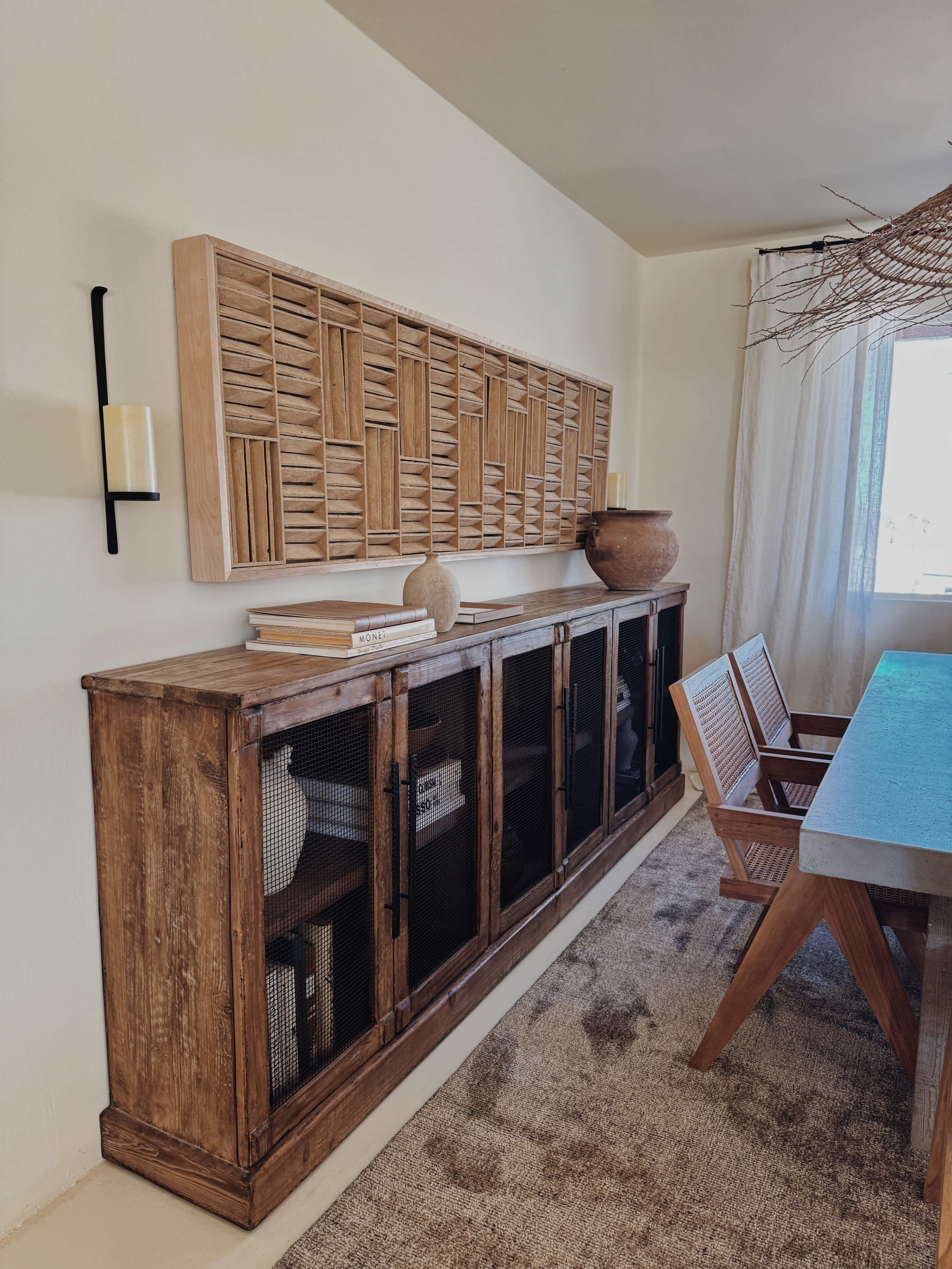 The image shows a wooden cabinet with glass doors displaying books and decor items, accompanied by a decorative wall panel and a dining table with wooden chairs in a room lit by a window.