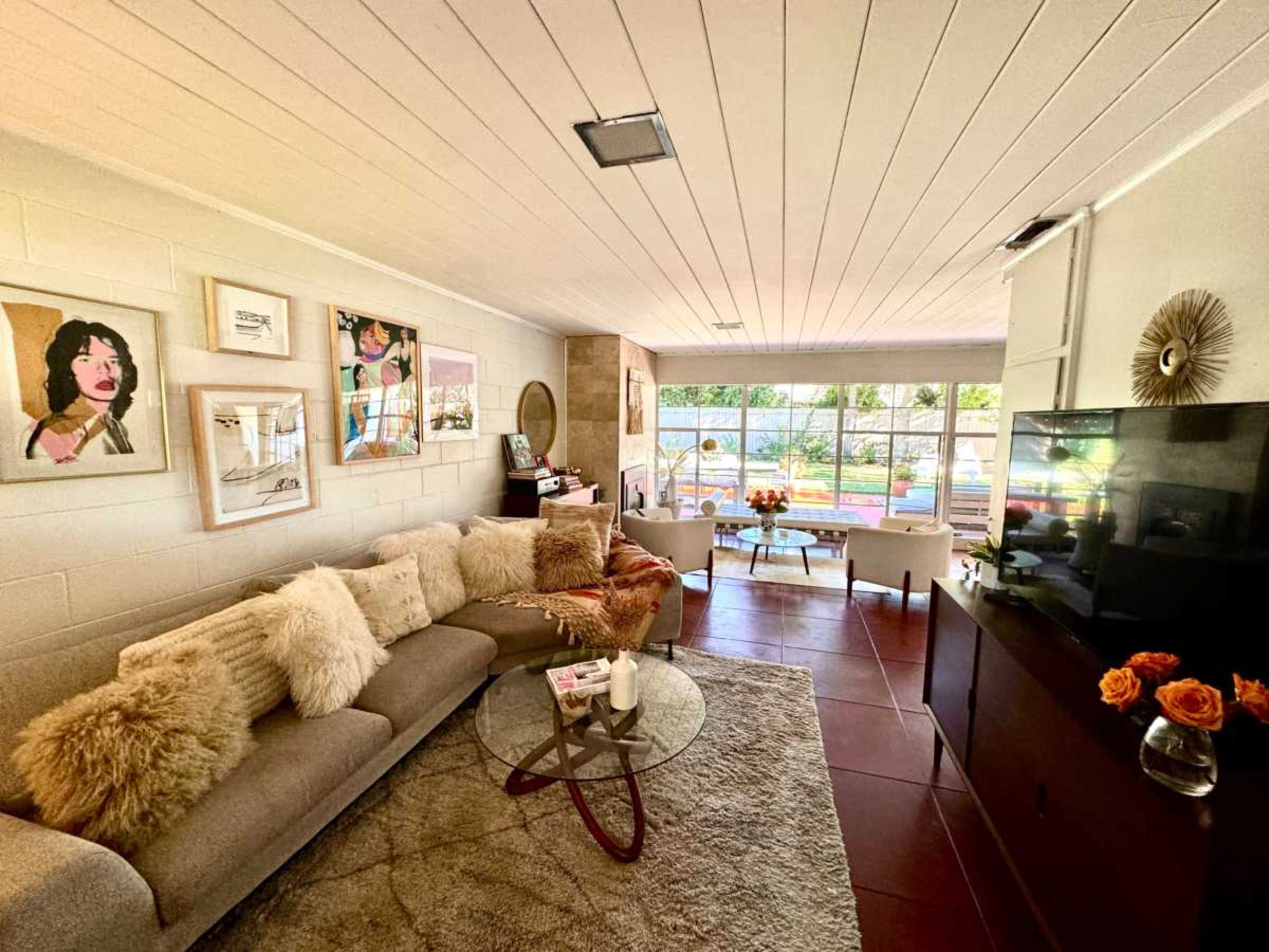 A modern living room features a large sectional sofa, a glass coffee table, and a view of a backyard through large windows.