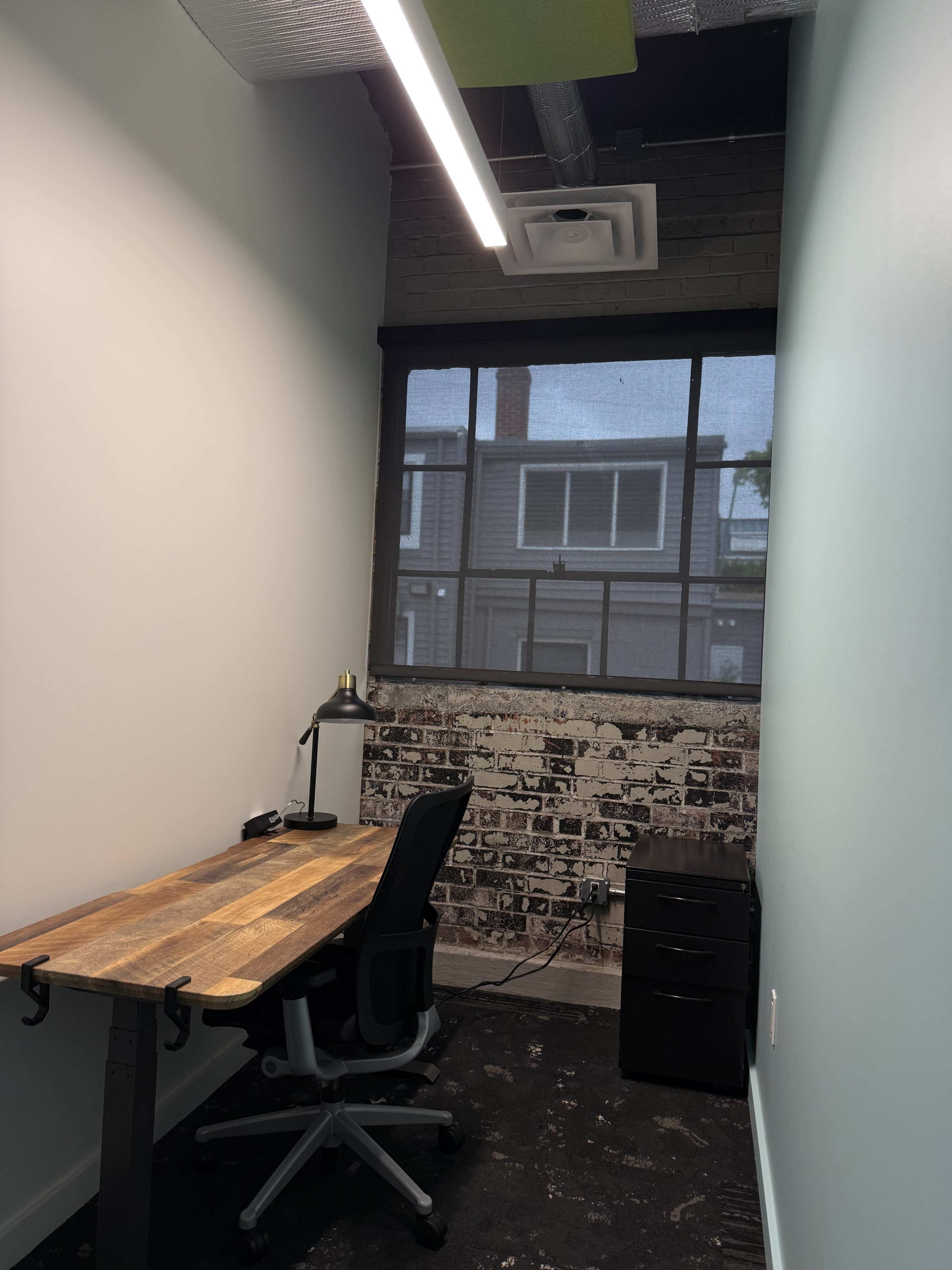 The image shows a small office space featuring a wooden desk, an ergonomic chair, and a black filing cabinet, with a large window revealing a view of a brick wall outside.