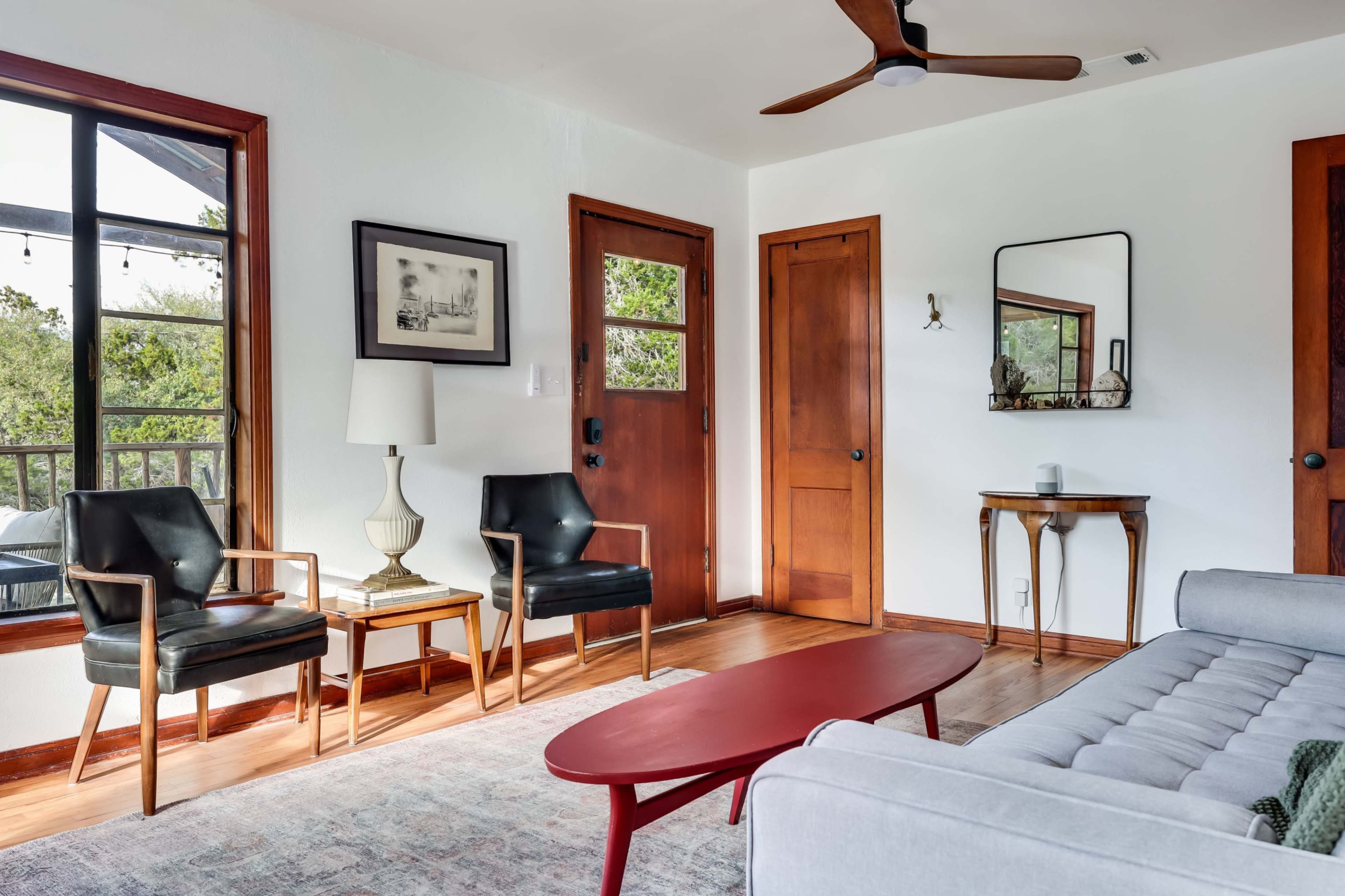 The image depicts a living room featuring two black chairs and a red coffee table, with wooden doors and a large window allowing natural light to enter the space.