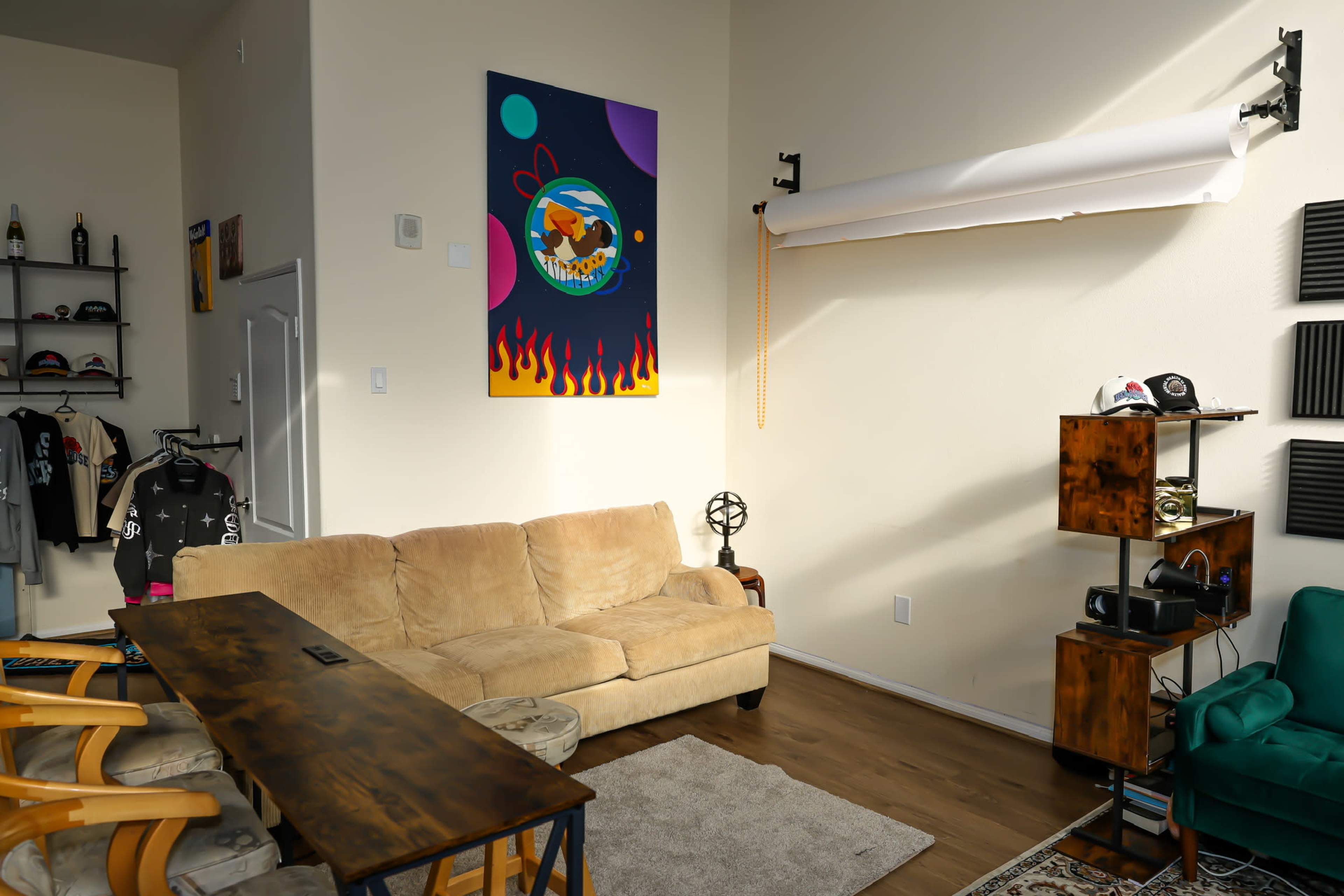 The image shows a cozy living space with a beige couch, a wooden coffee table, and a vibrant wall art piece featuring an abstract design, alongside shelves displaying various clothing items and accessories.