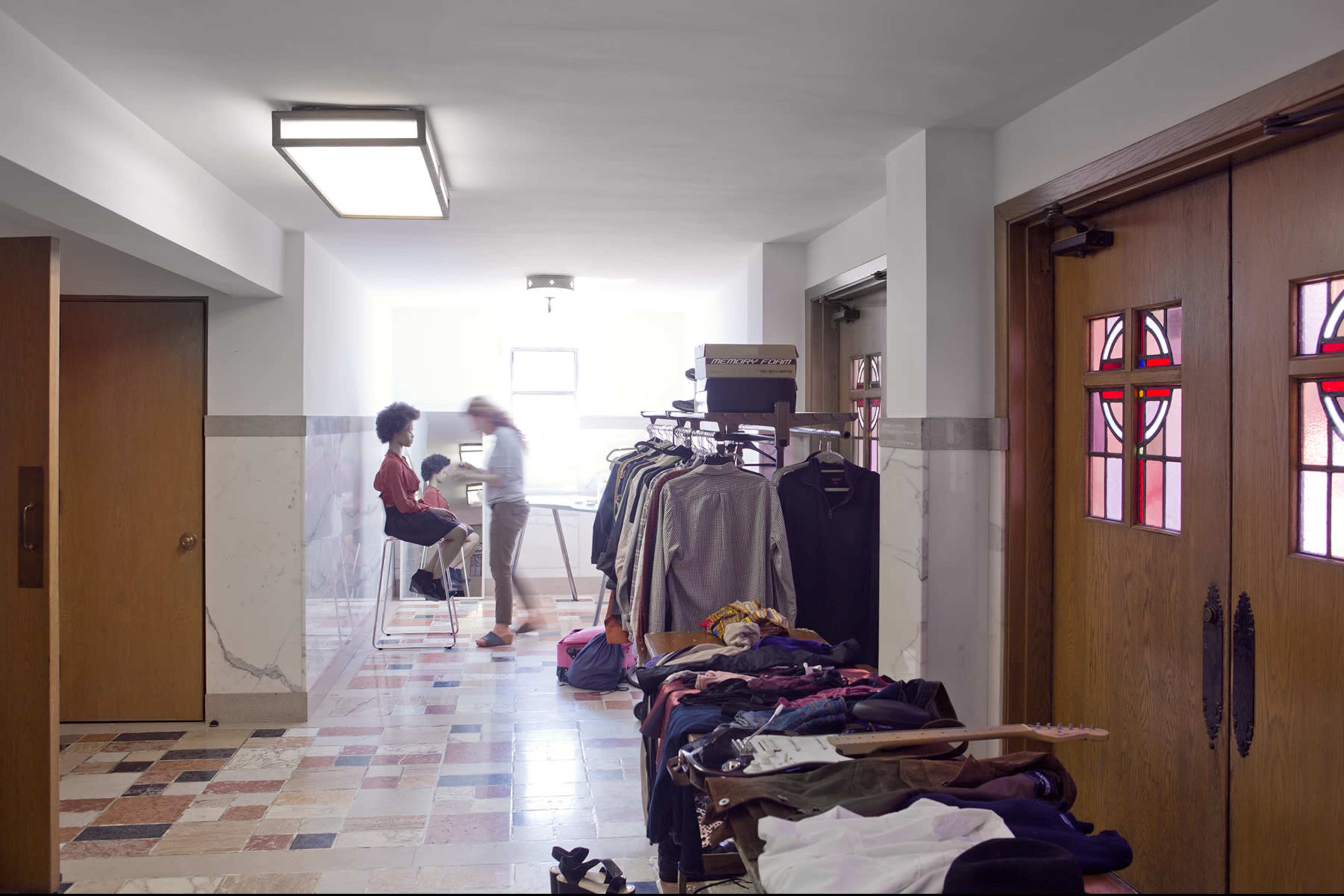 A hallway features a clothing rack filled with garments on one side, while two people are engaged in conversation near a window.