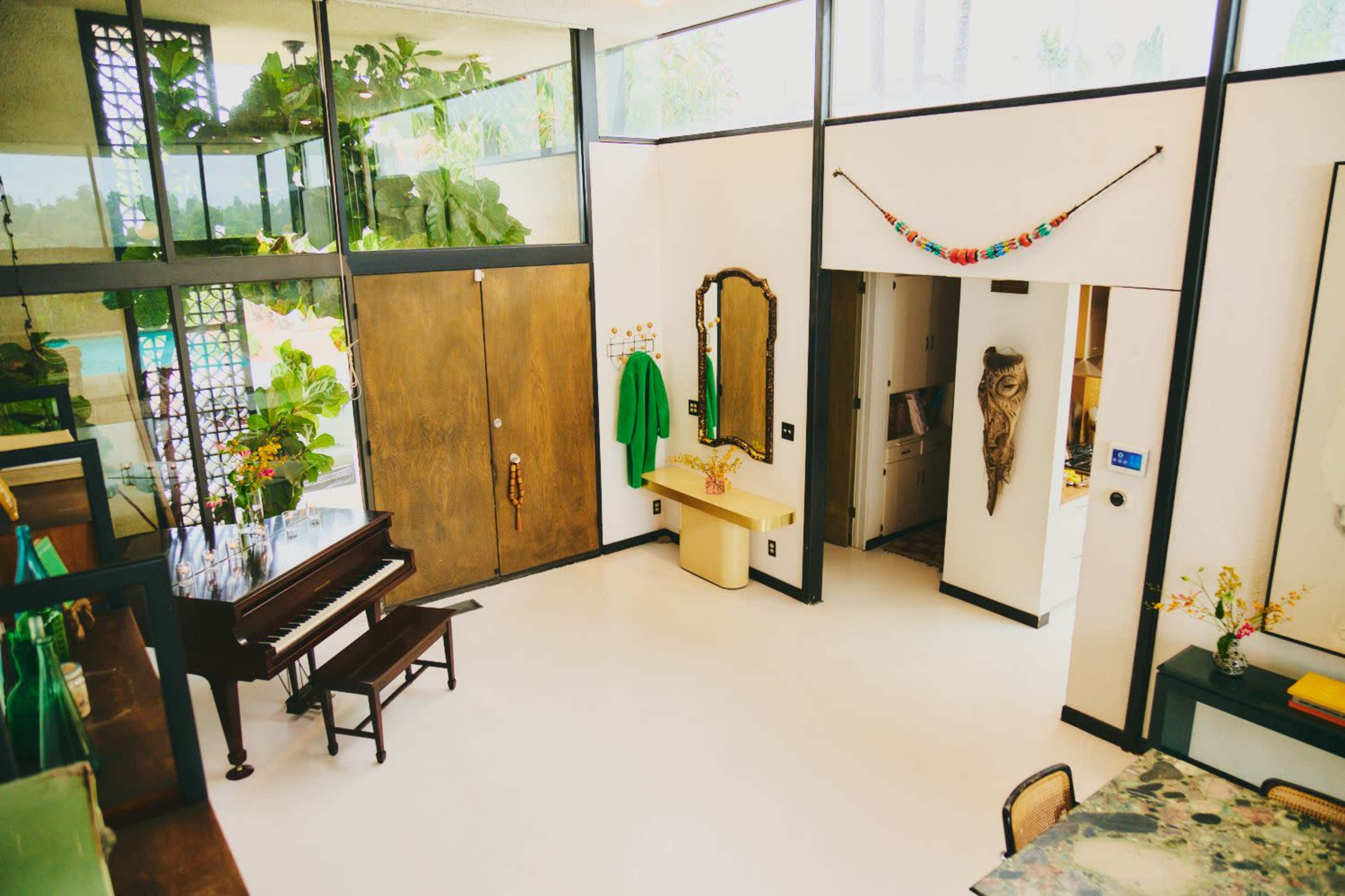 The image shows a modern interior space featuring a piano, a yellow console table, large windows, and decorative greenery.