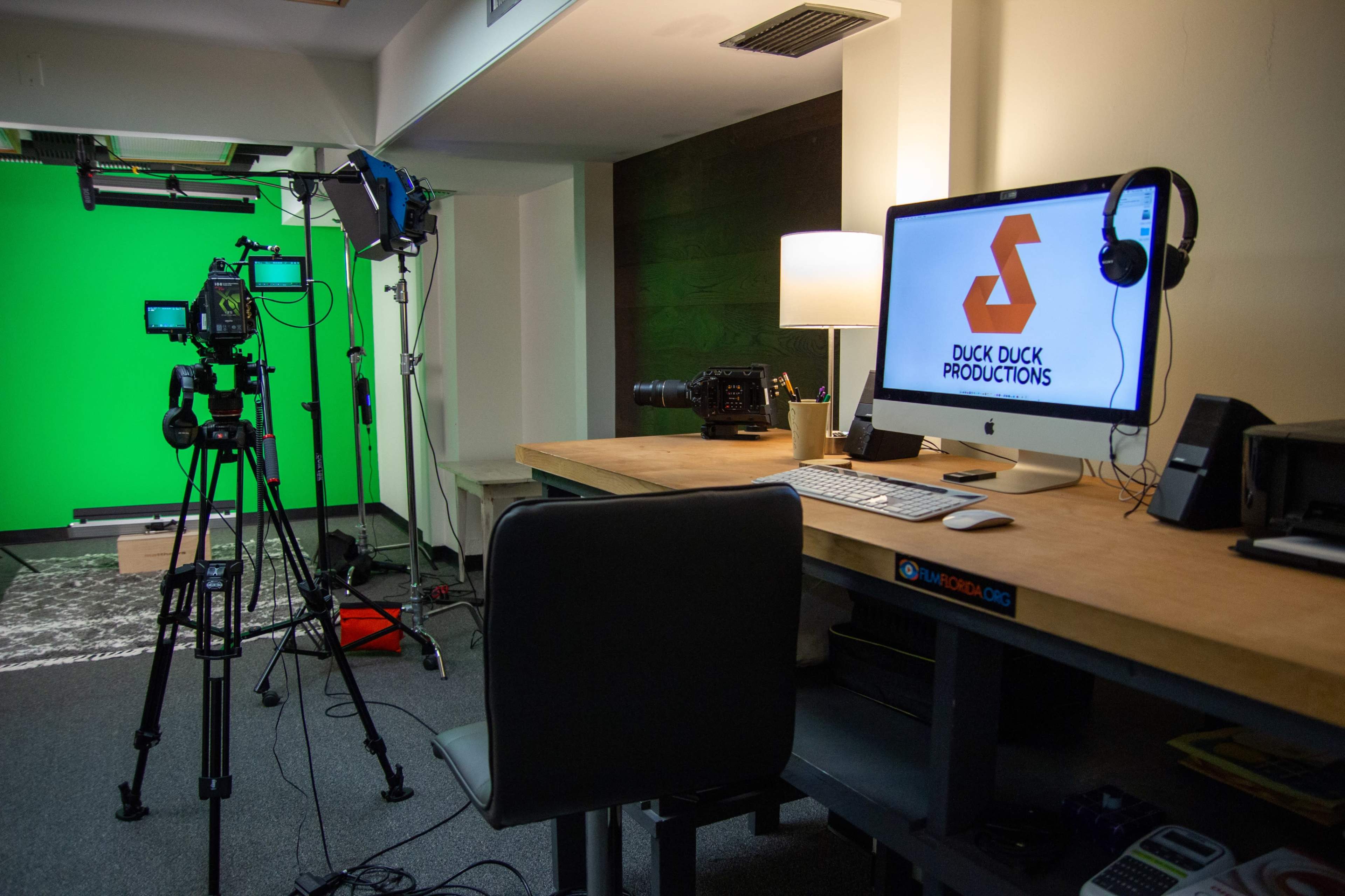 A production studio with a green screen, camera equipment, a computer displaying the logo "DUCK DUCK PRODUCTIONS," and a desk with a chair.