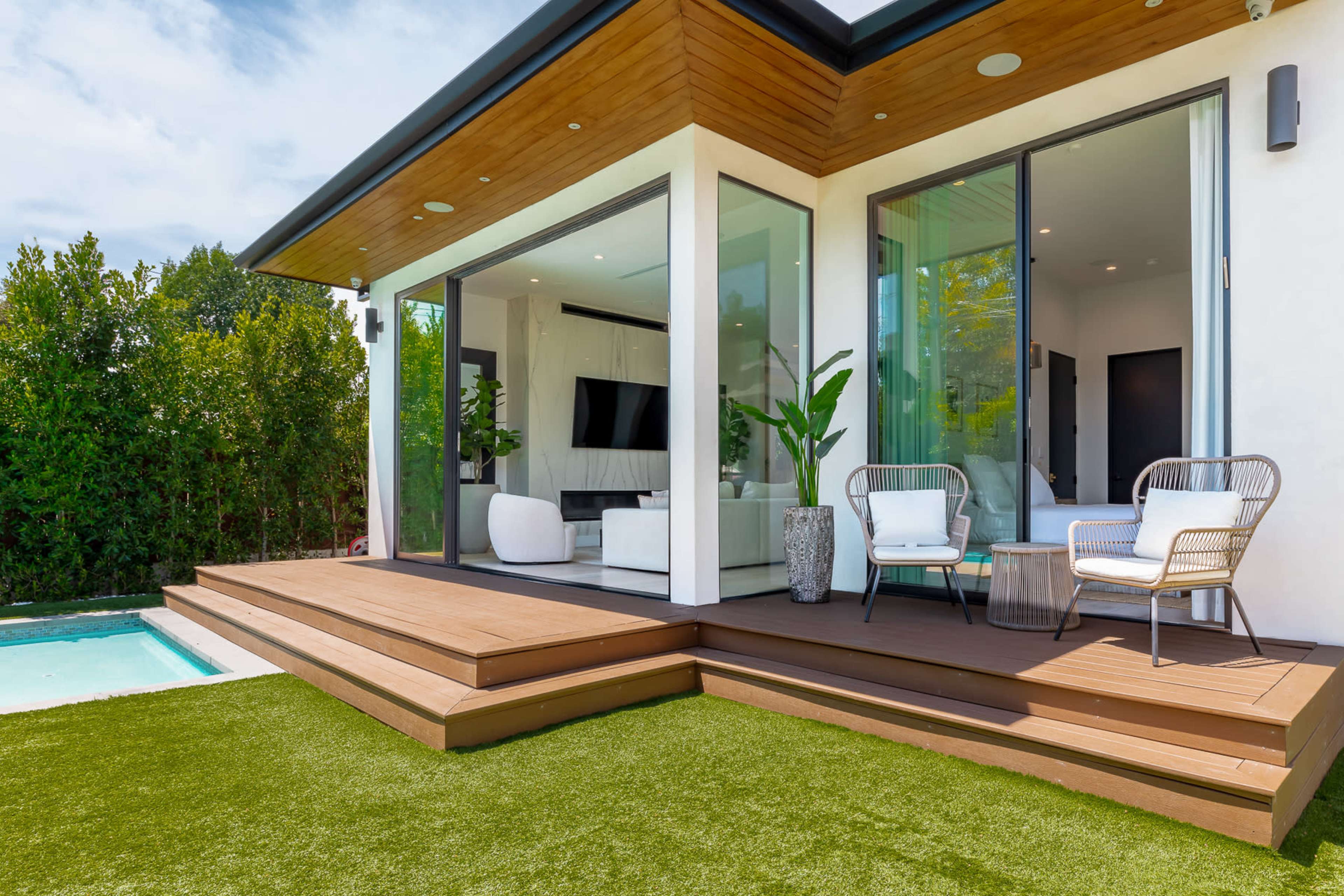 A modern house features a spacious deck with seating areas, surrounded by lush greenery and a swimming pool.