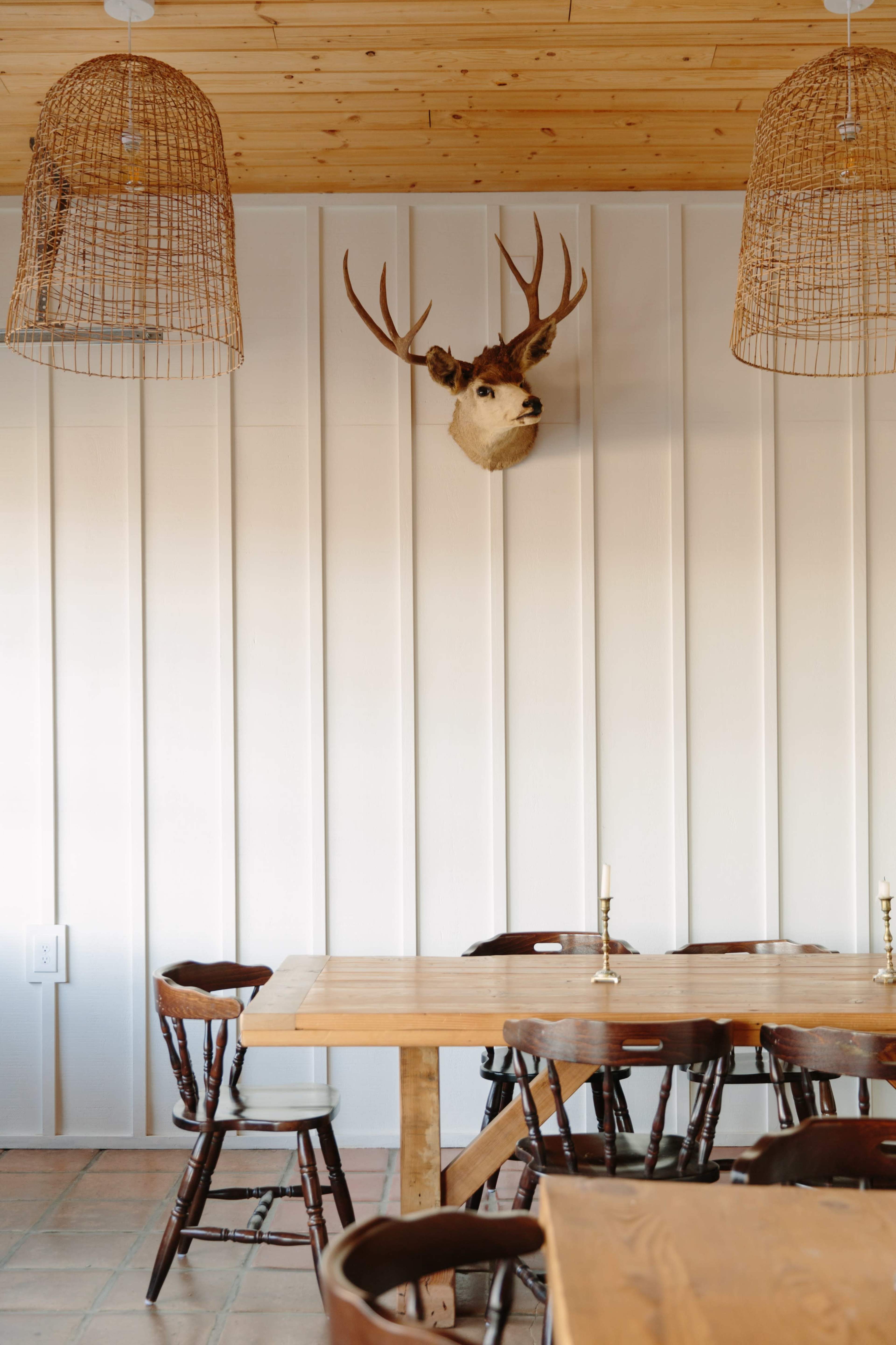 A wooden table with chairs sits beneath two woven light fixtures, while a mounted deer head adorns the wall behind it.
