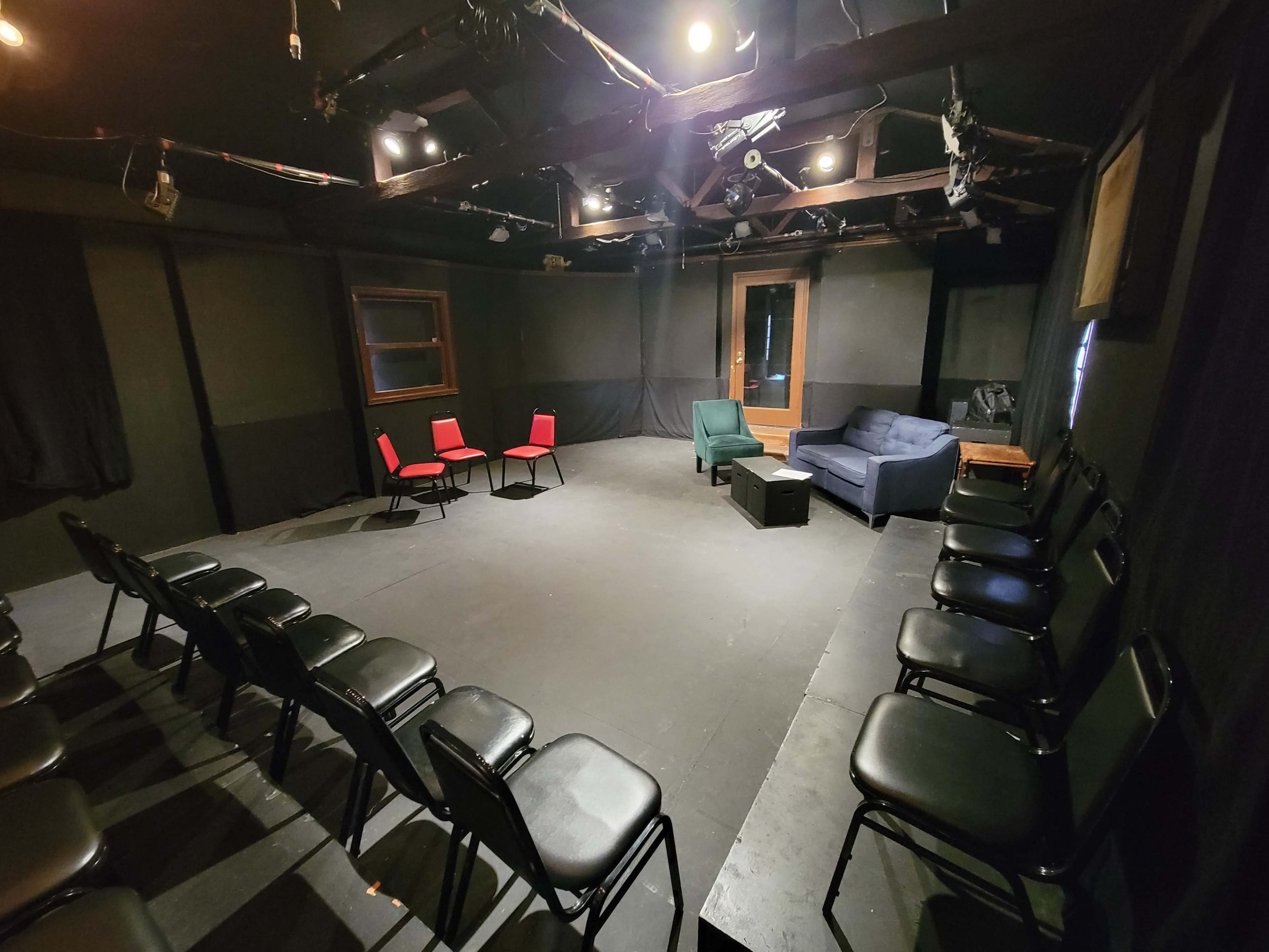 The image shows an empty theater rehearsal space with black walls, a wooden beam ceiling, and rows of black chairs facing a central seating area that includes a green chair and a blue couch.