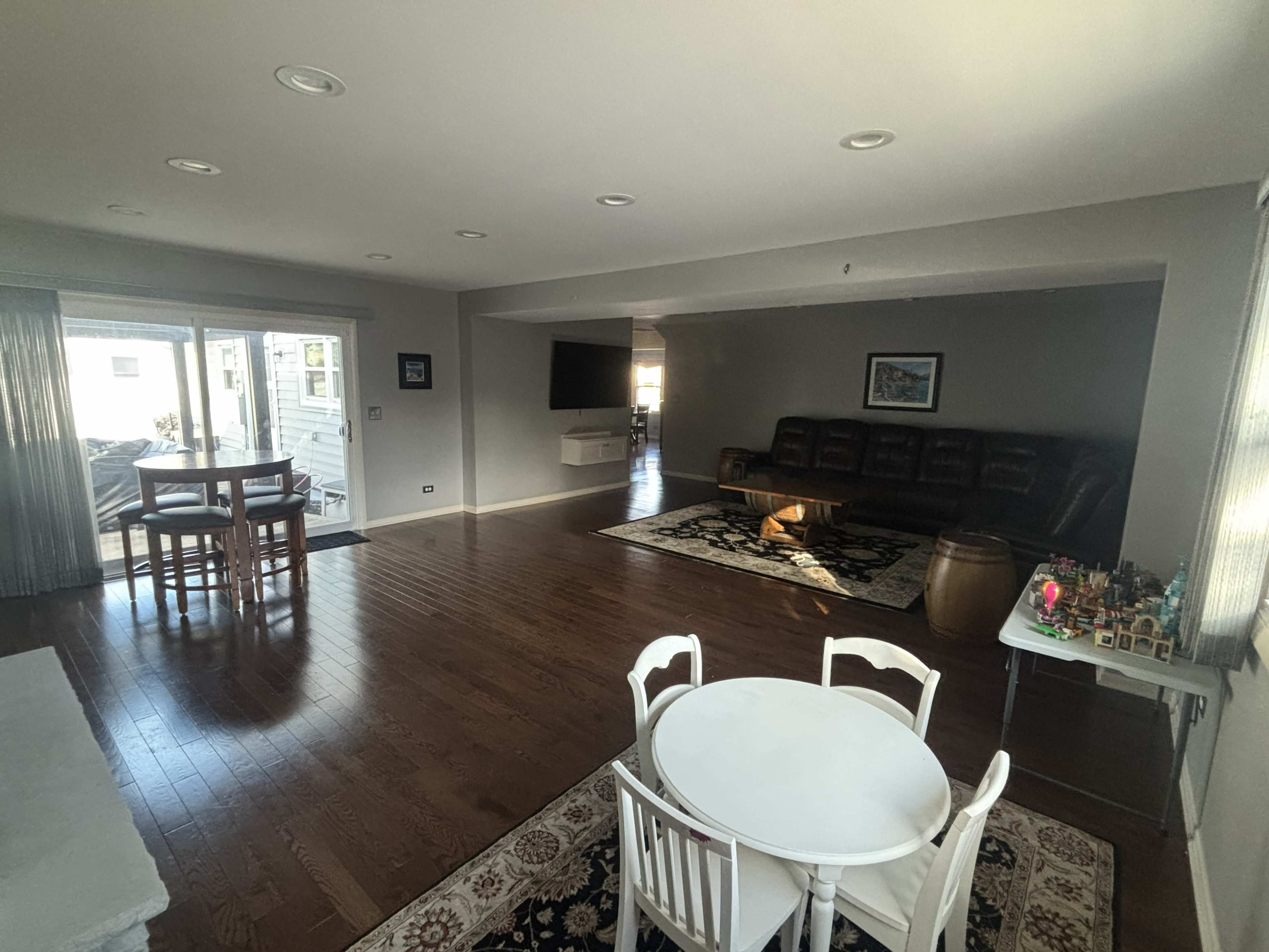 The image shows a spacious living area with hardwood floors, a dark leather sofa, a small dining table, and large windows allowing natural light.