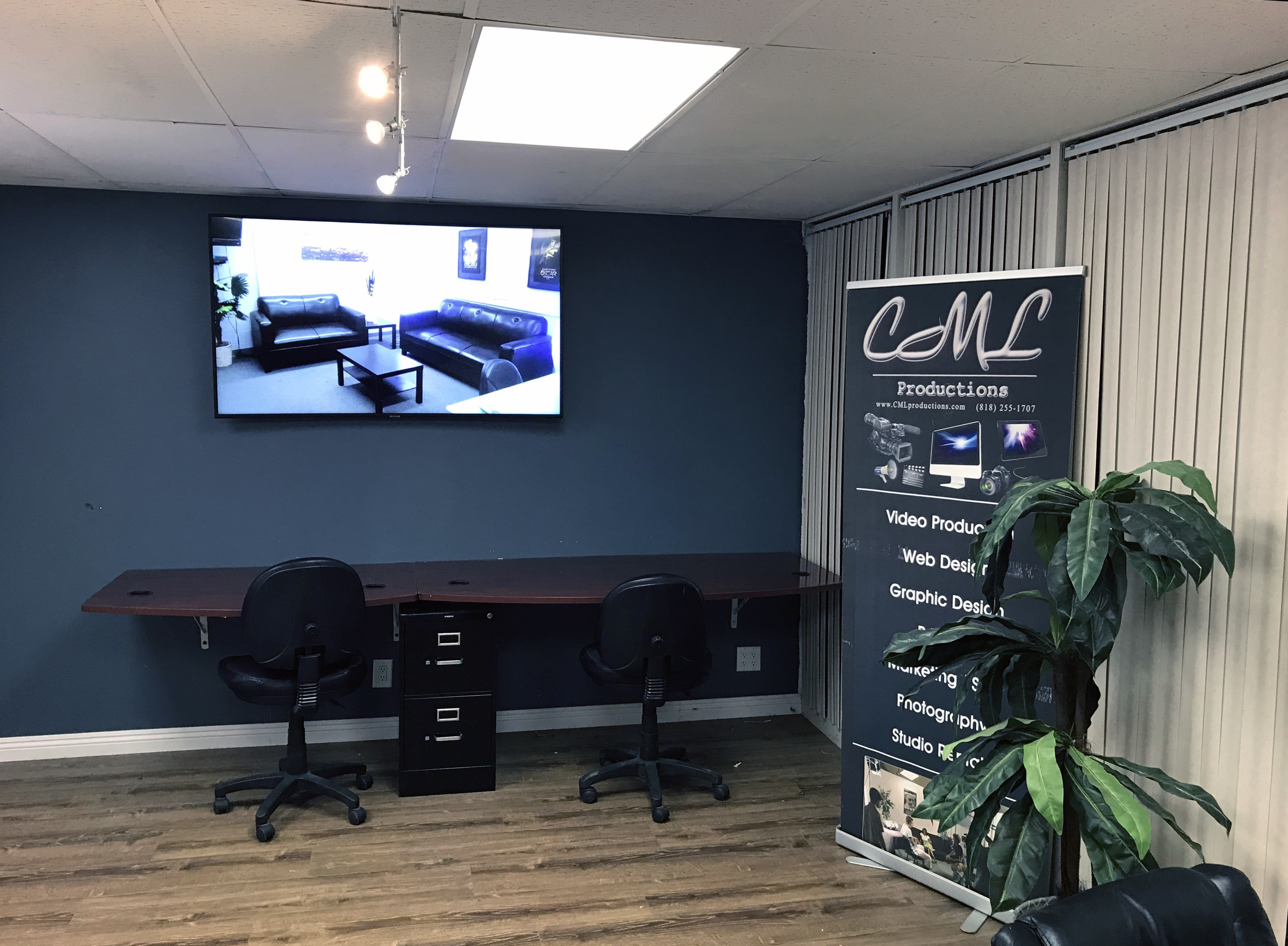 An office space with a large television on the wall, a desk with two black chairs, a potted plant, and a banner for a production company.