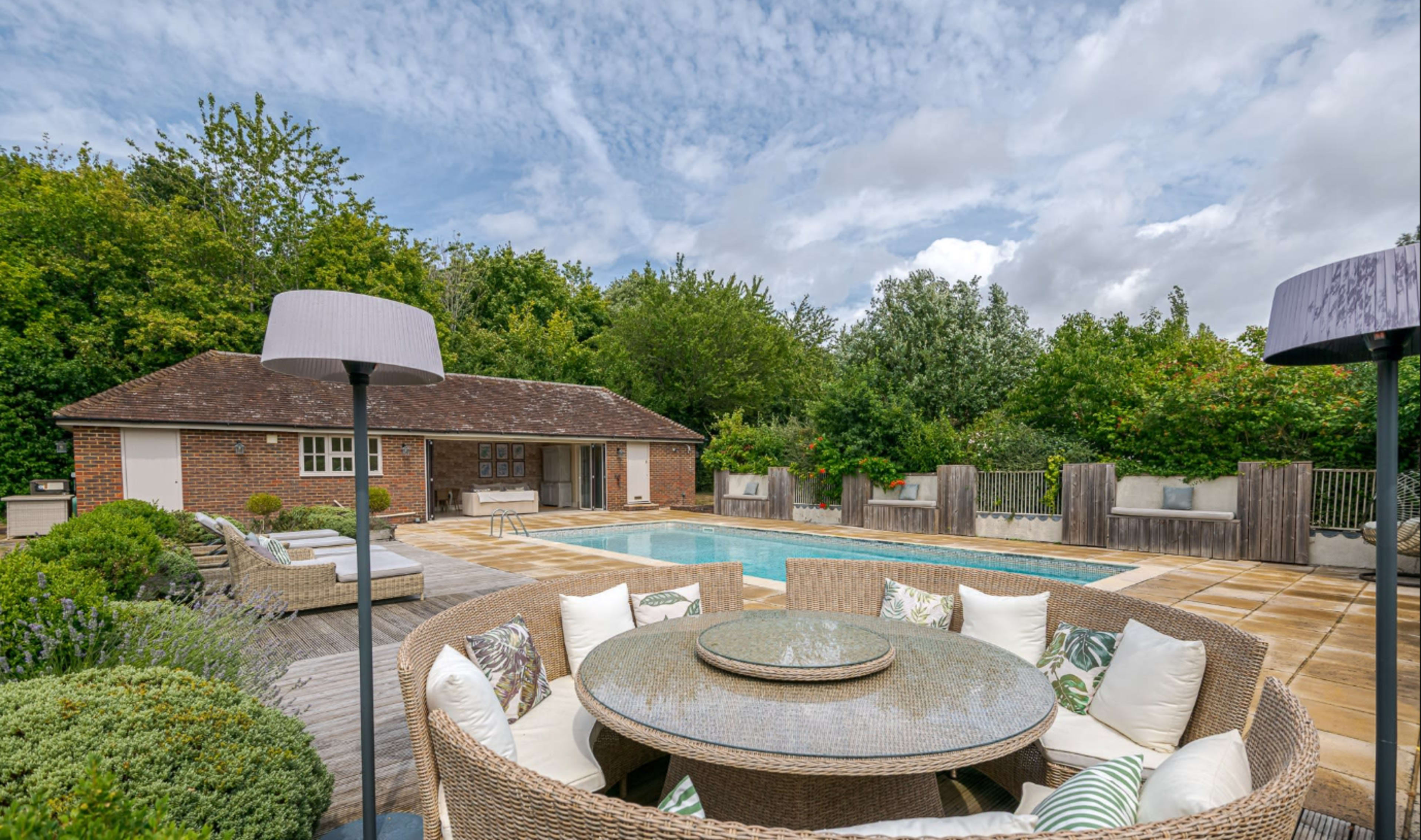 The image shows a backyard pool area with a circular dining table surrounded by comfortable seating, and a brick building and lush greenery in the background.