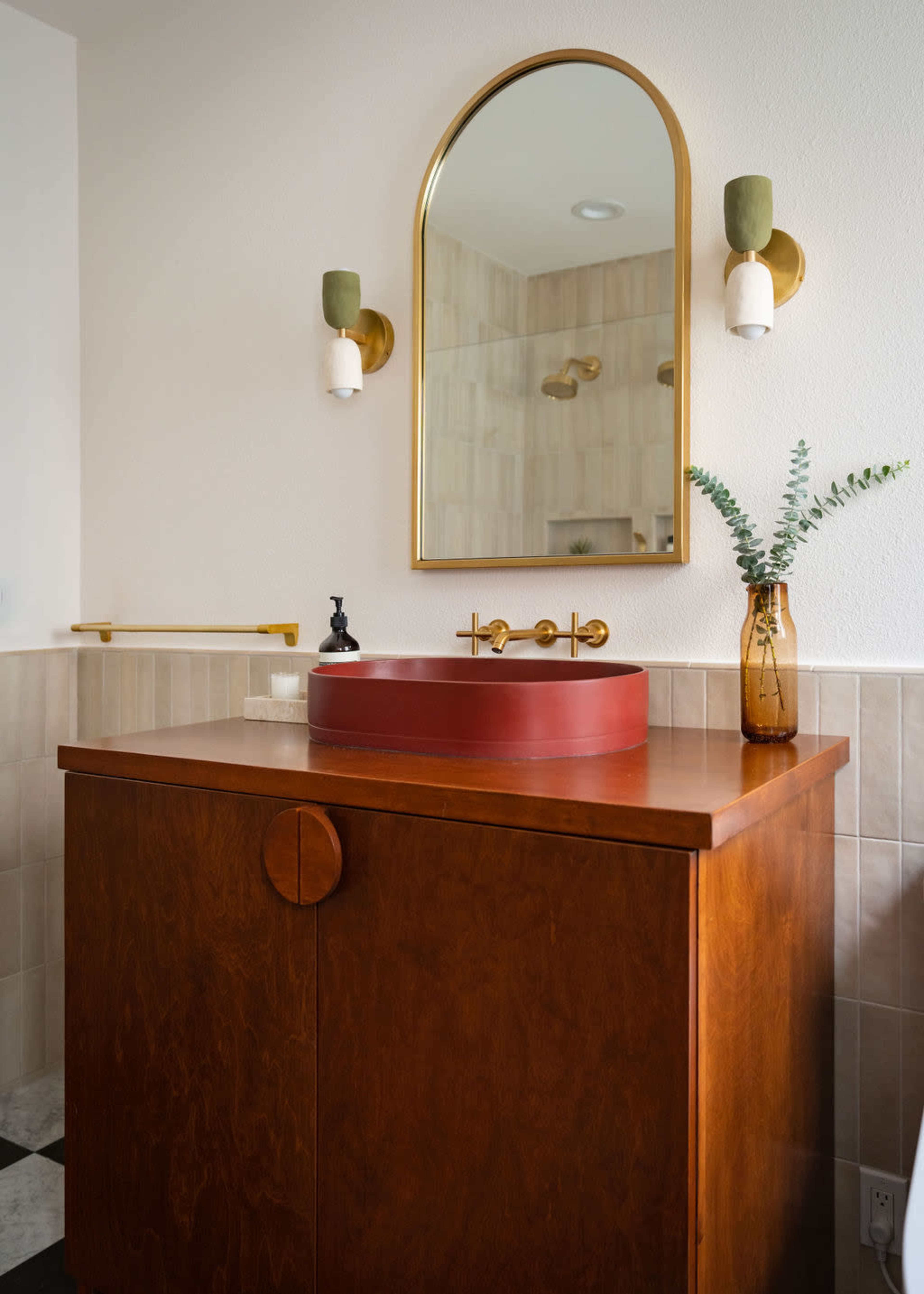 A modern bathroom features a wooden vanity with a round red sink, a wall mirror with a gold frame, and stylish sconces.