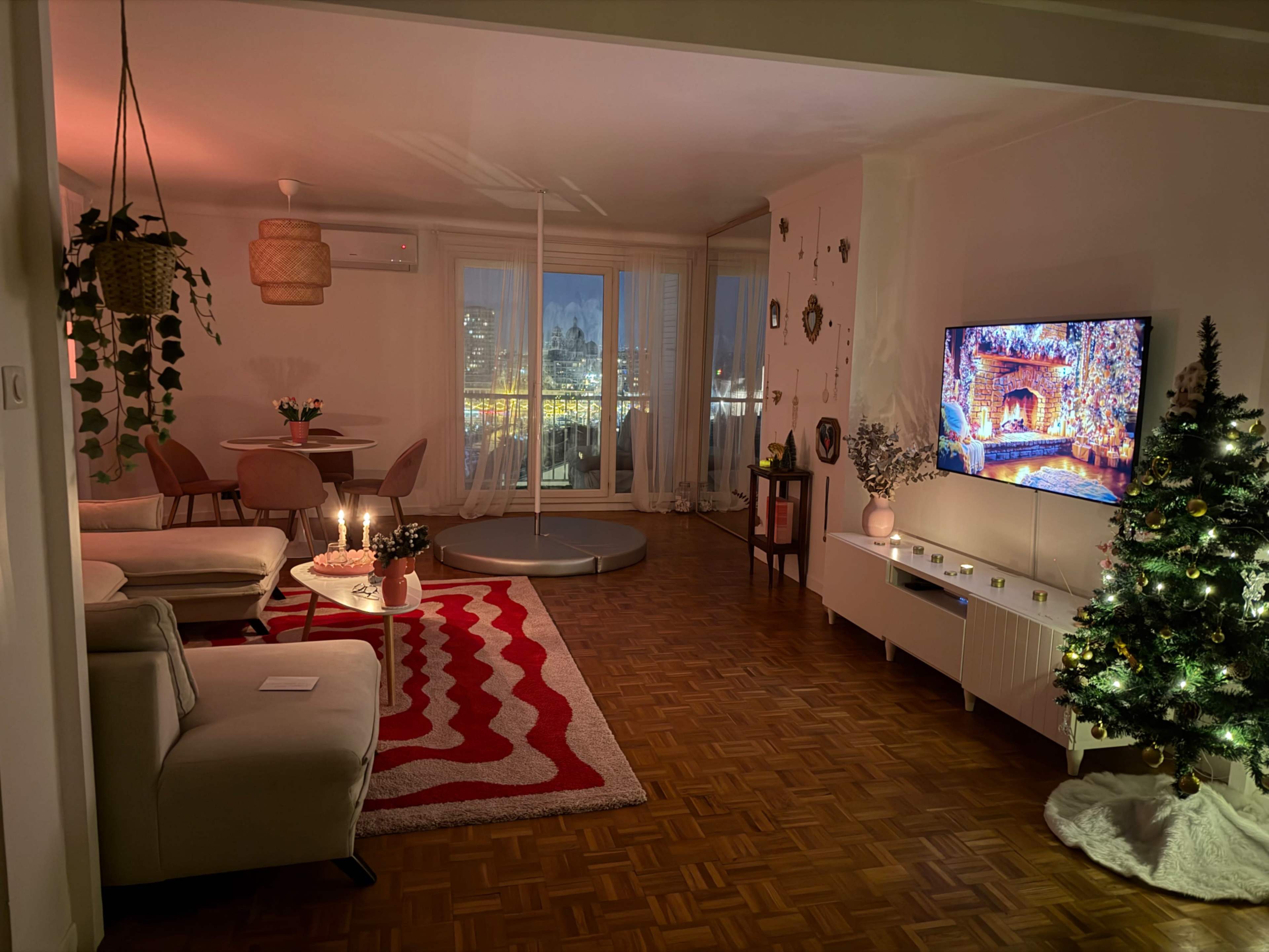 The image shows a cozy living room with a Christmas tree, a small dining area, and a TV displaying a festive scene, all illuminated by soft lighting.
