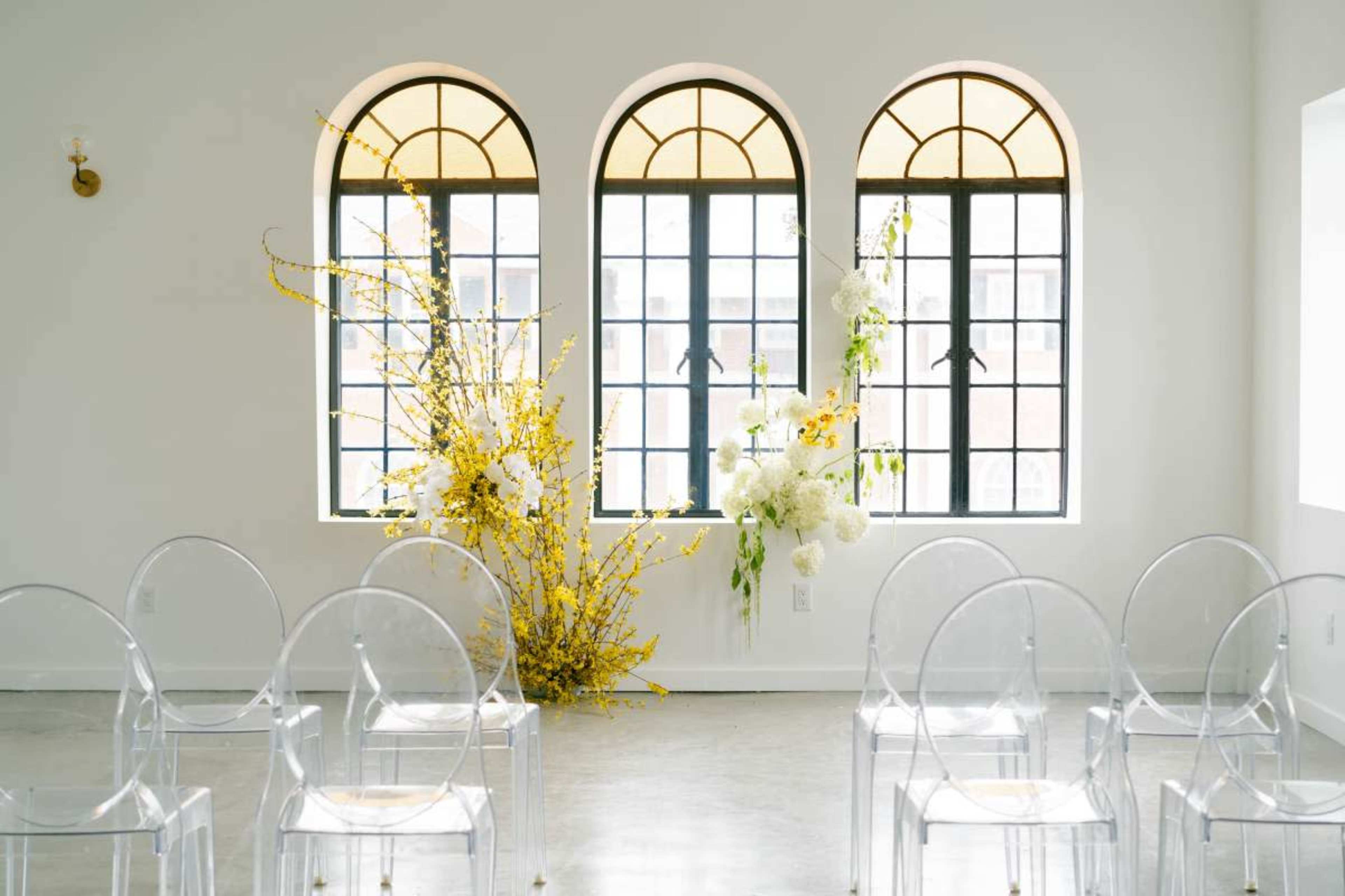 The image shows a bright, minimalist room with three tall windows, two floral arrangements on either side, and transparent chairs arranged in front.