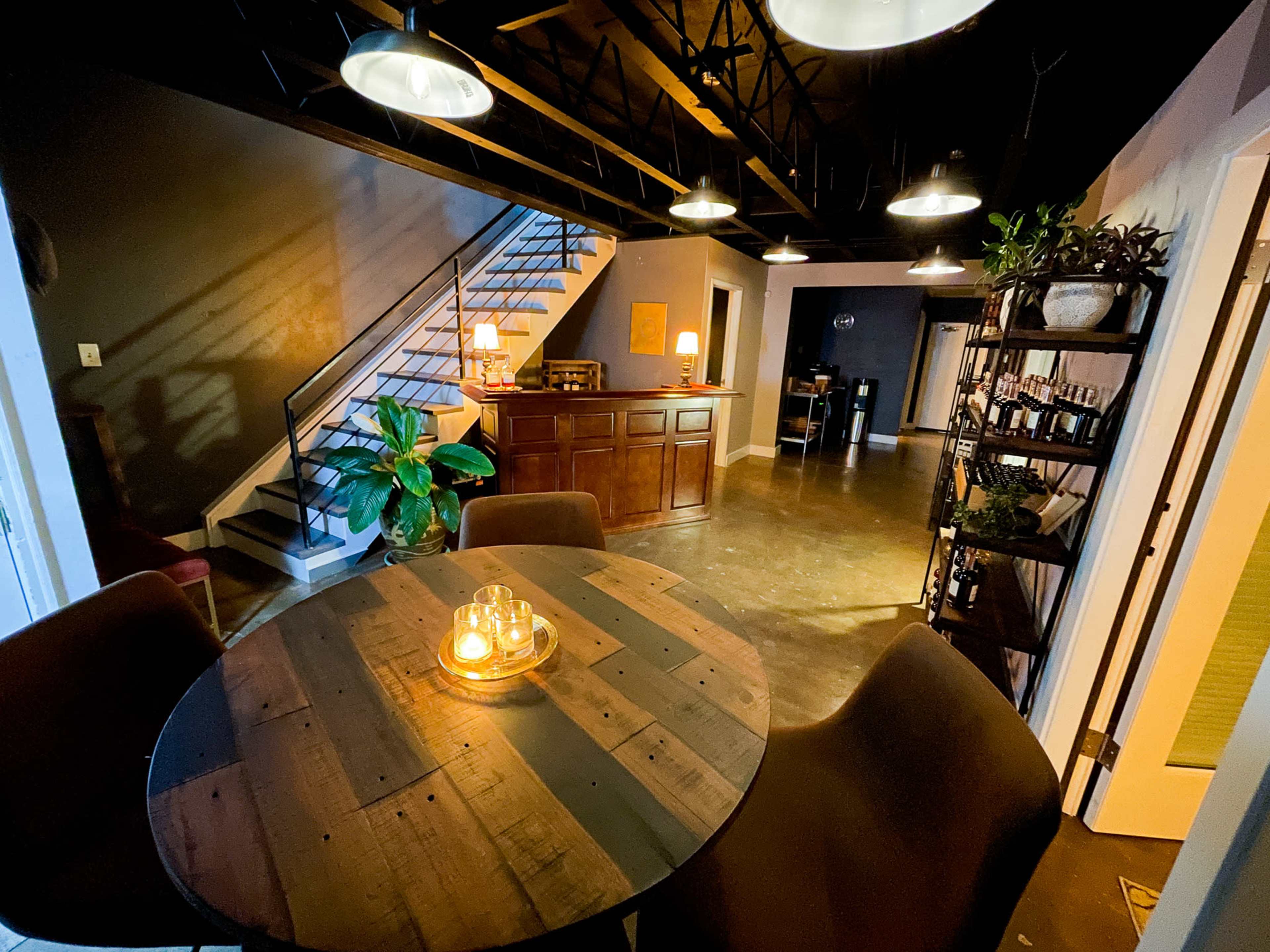 The image shows a spacious, modern interior with a round wooden table at the center, surrounded by chairs, and a staircase leading to an upper level.