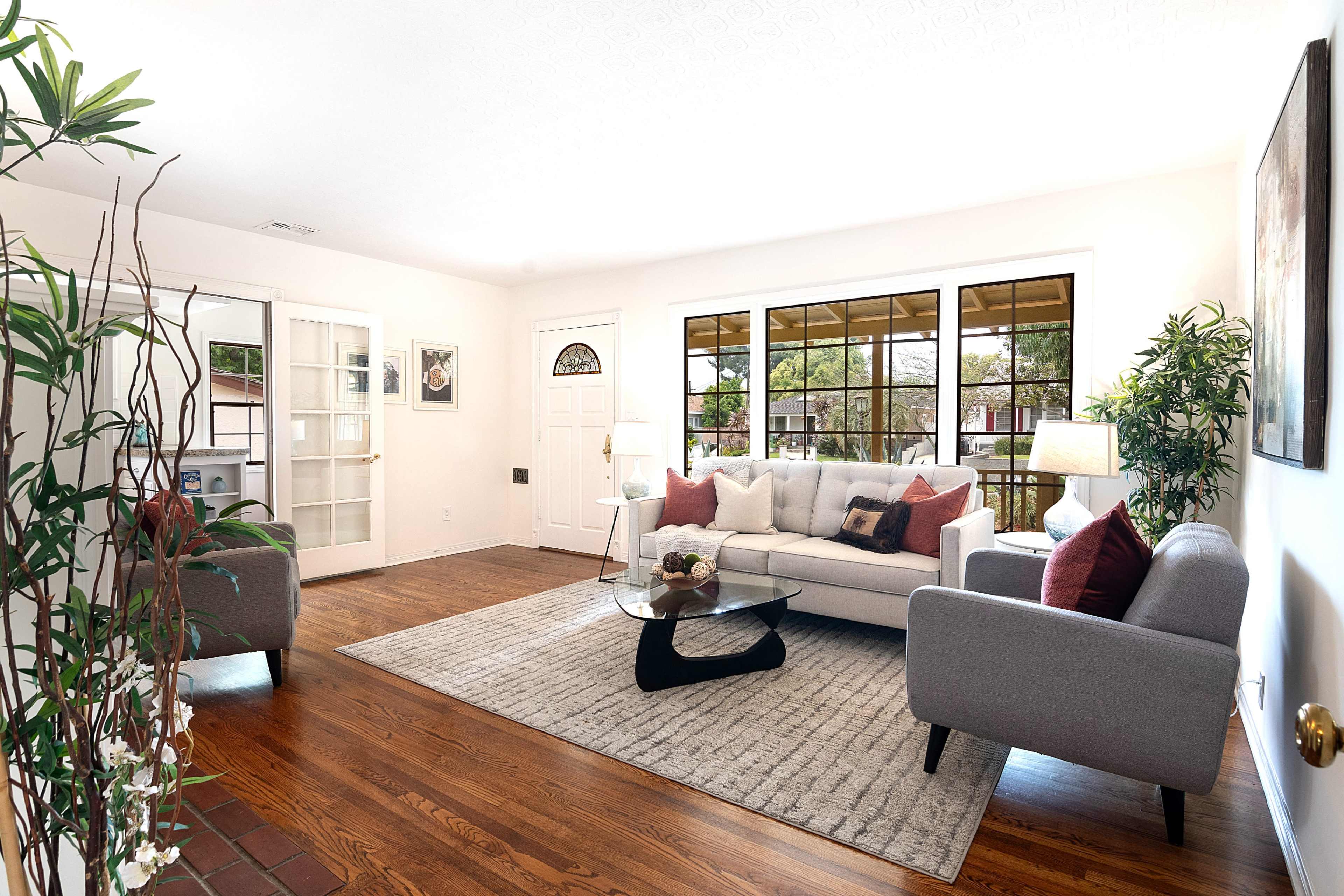 A well-lit living room features a gray sofa with red accent pillows, a glass coffee table, and large windows that open to a view of greenery outside.