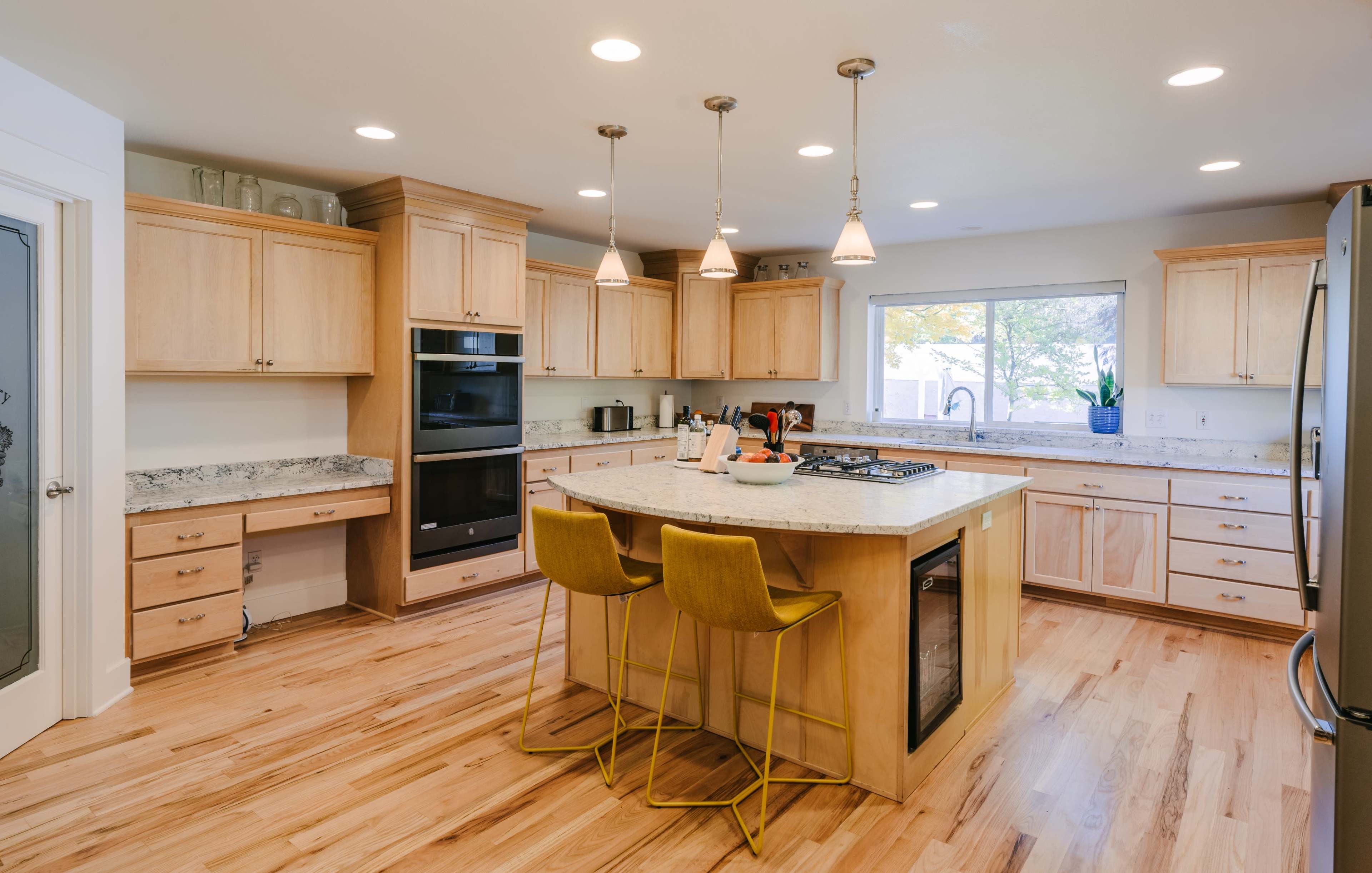 A modern kitchen features light wood cabinets, a central island with three yellow bar stools, and stainless steel appliances.