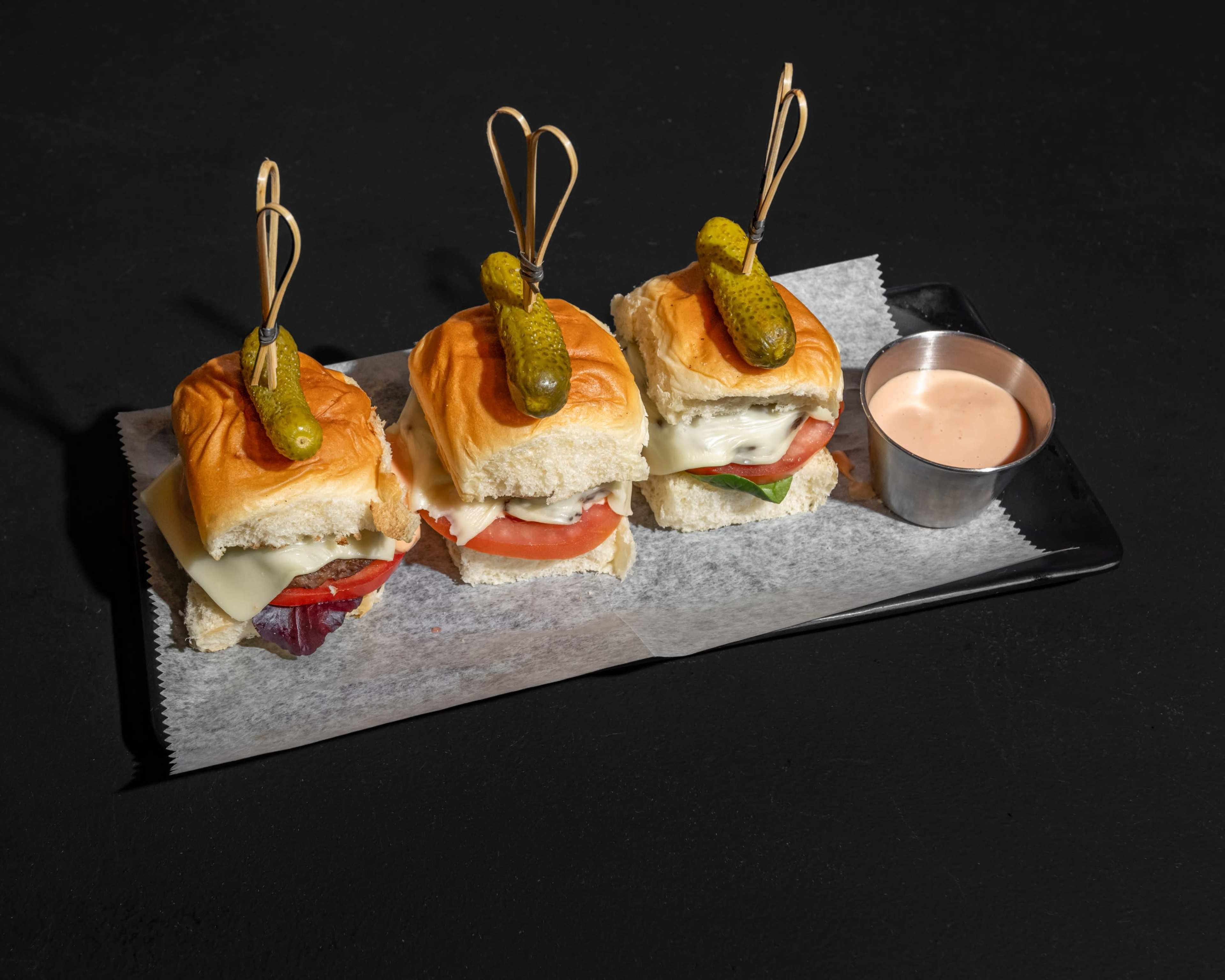 The image displays three small sandwiches on a black plate, each topped with a pickle and accompanied by a small cup of sauce.