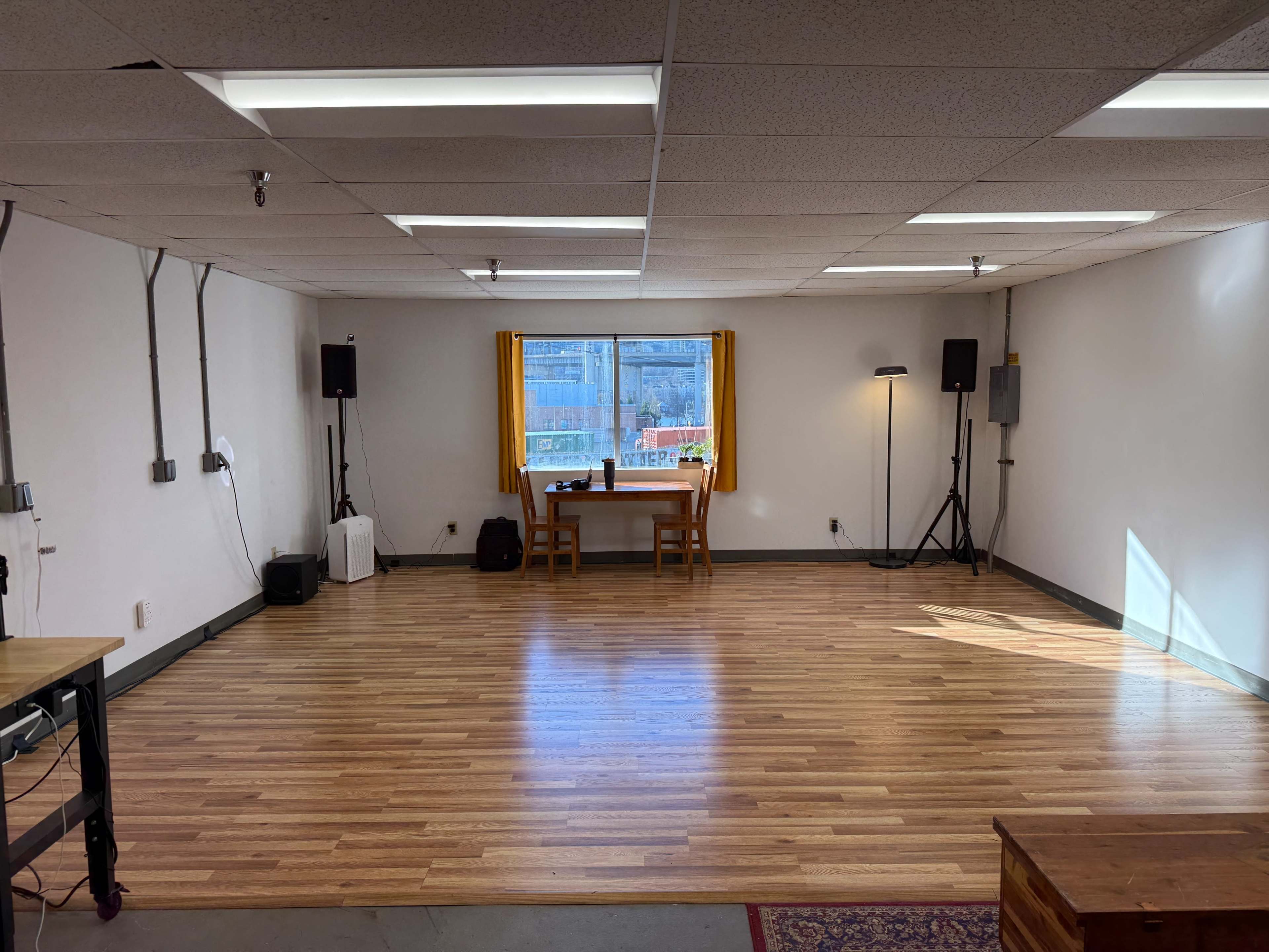 The image shows a spacious, empty room with wooden flooring, large windows with yellow curtains, and a table in the center flanked by audio equipment on either side.