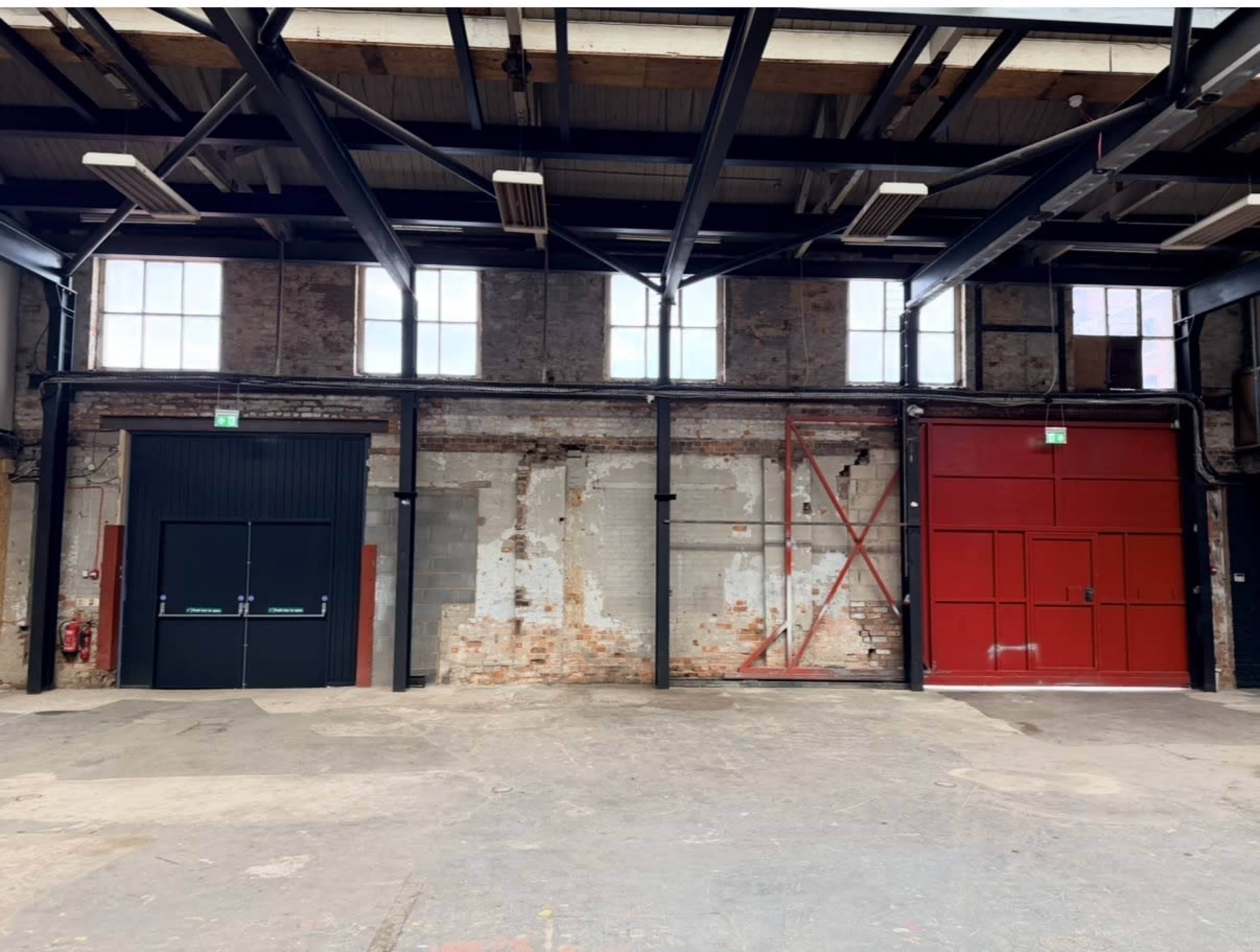 The image shows a spacious industrial interior featuring two large doors—one black and the other red—set against a brick wall with exposed beams and large windows above.