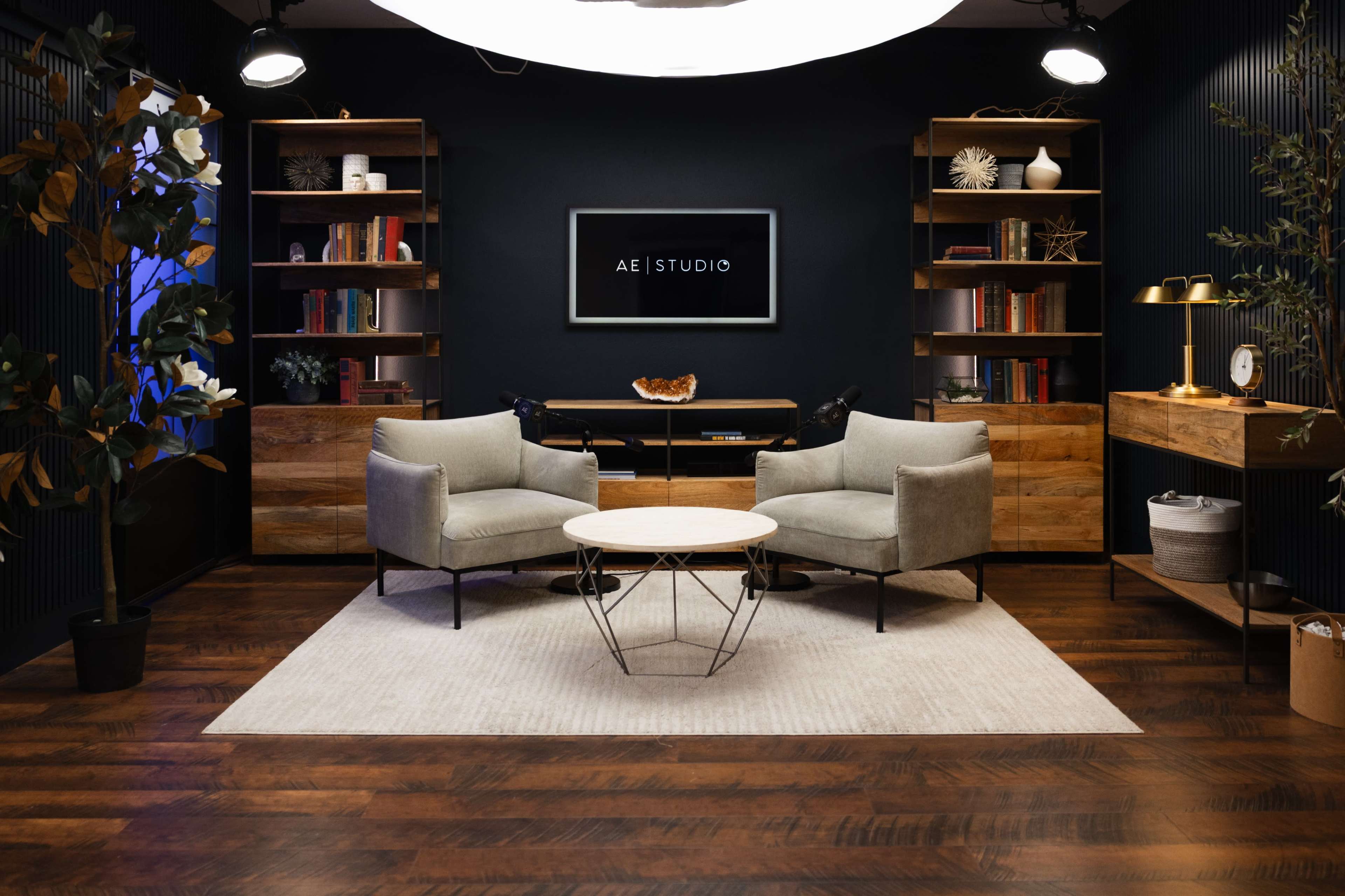 A modern studio setup features two gray armchairs facing a round coffee table, surrounded by wooden shelves filled with books and decor, all set against a dark background.