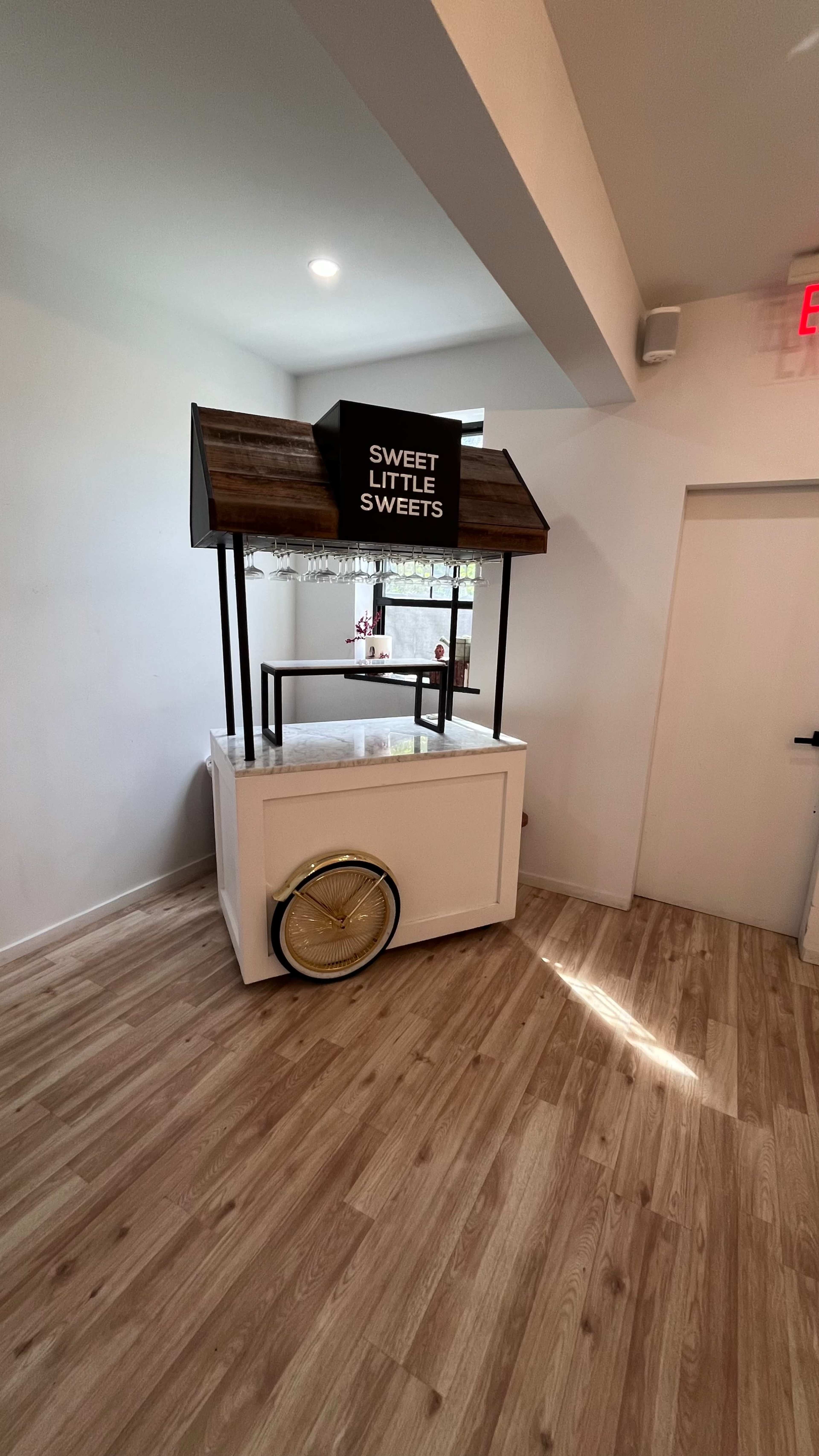 A small retail kiosk with a wooden roof labeled "SWEET LITTLE SWEETS" is positioned in a well-lit room with wooden flooring.