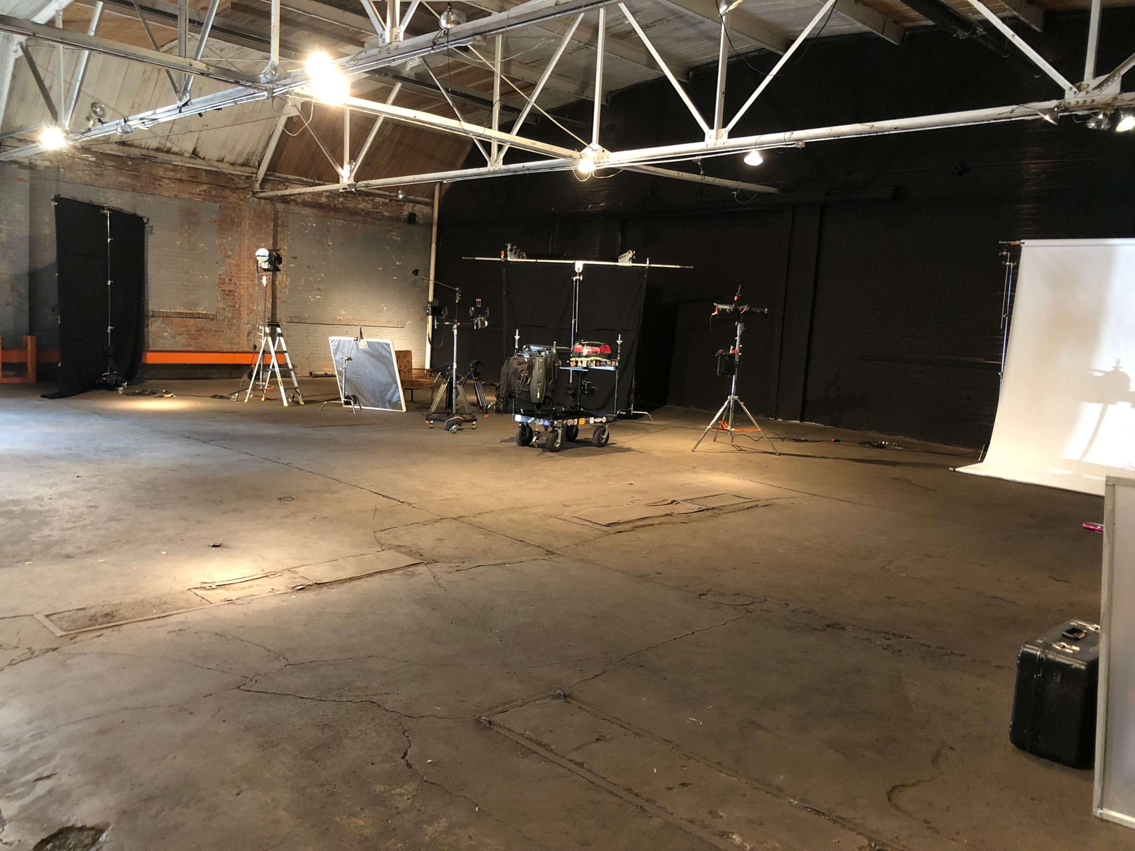 An empty industrial space with a concrete floor, featuring lighting equipment, a drum set, and black backdrops.