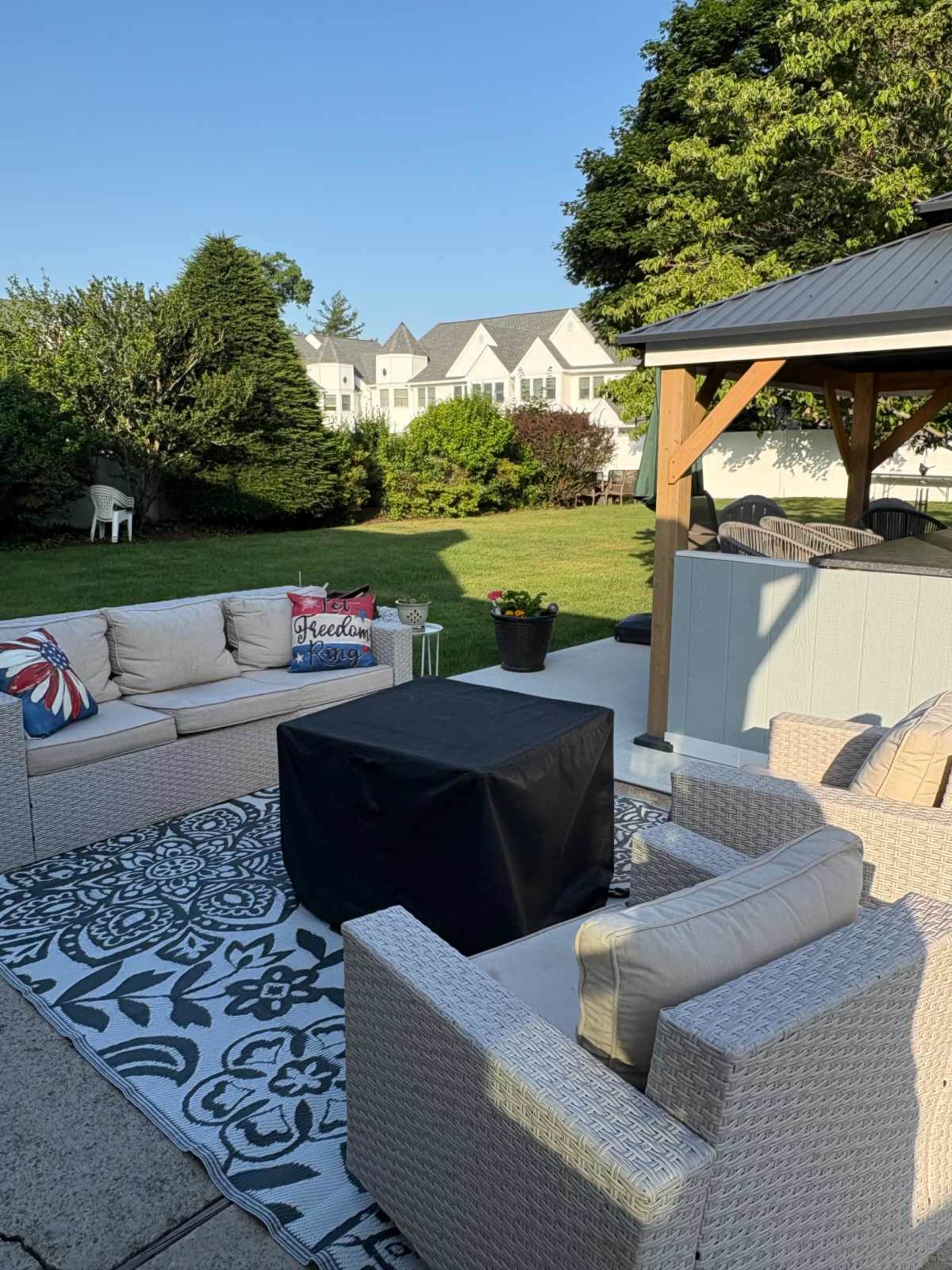 The image shows a landscaped backyard with a seating area, a covered gazebo, and a coffee table surrounded by light-colored outdoor furniture on a patterned rug.