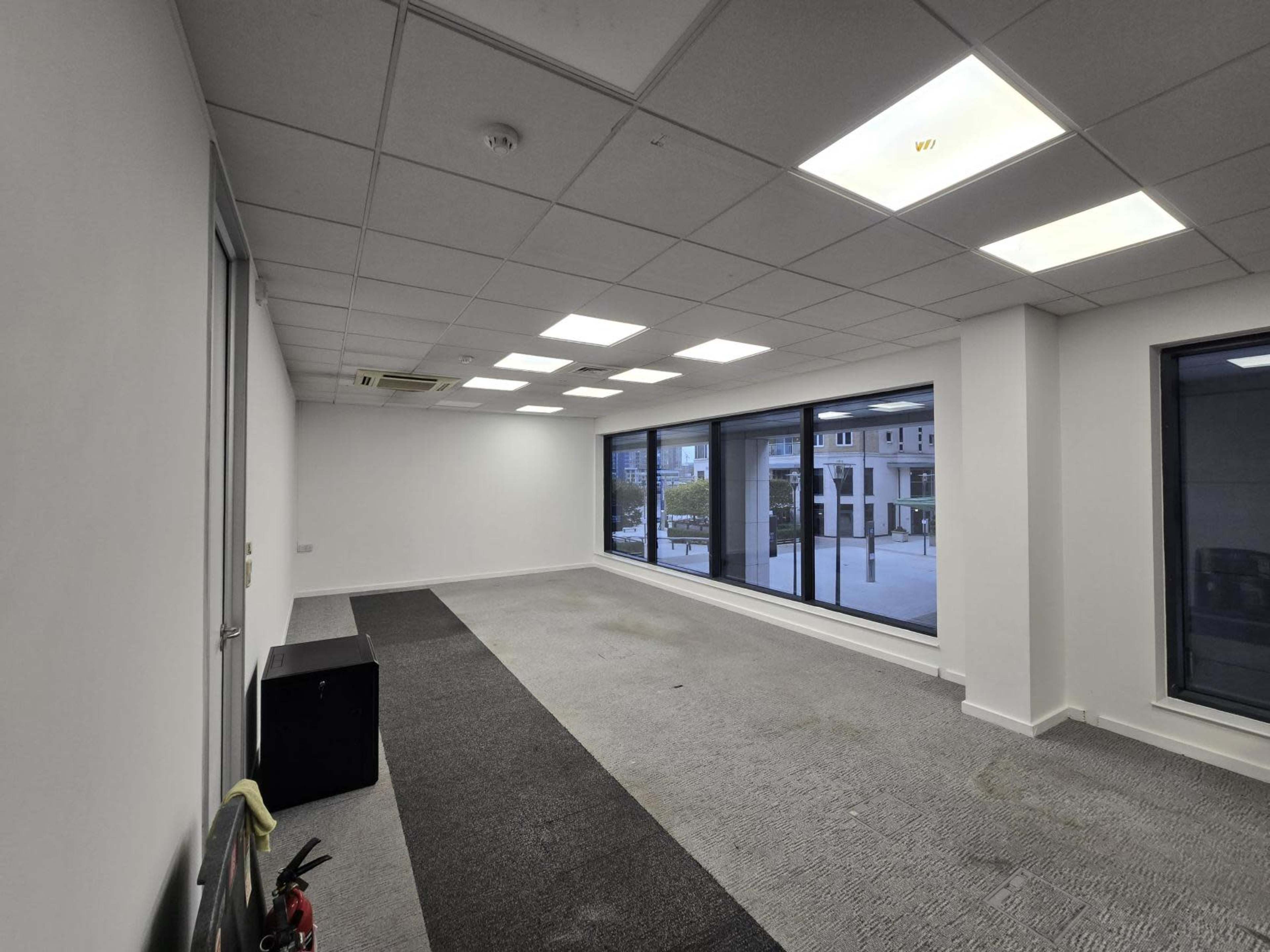 The image shows an empty office space with large windows and a carpeted floor, featuring a mix of gray and black carpeting.