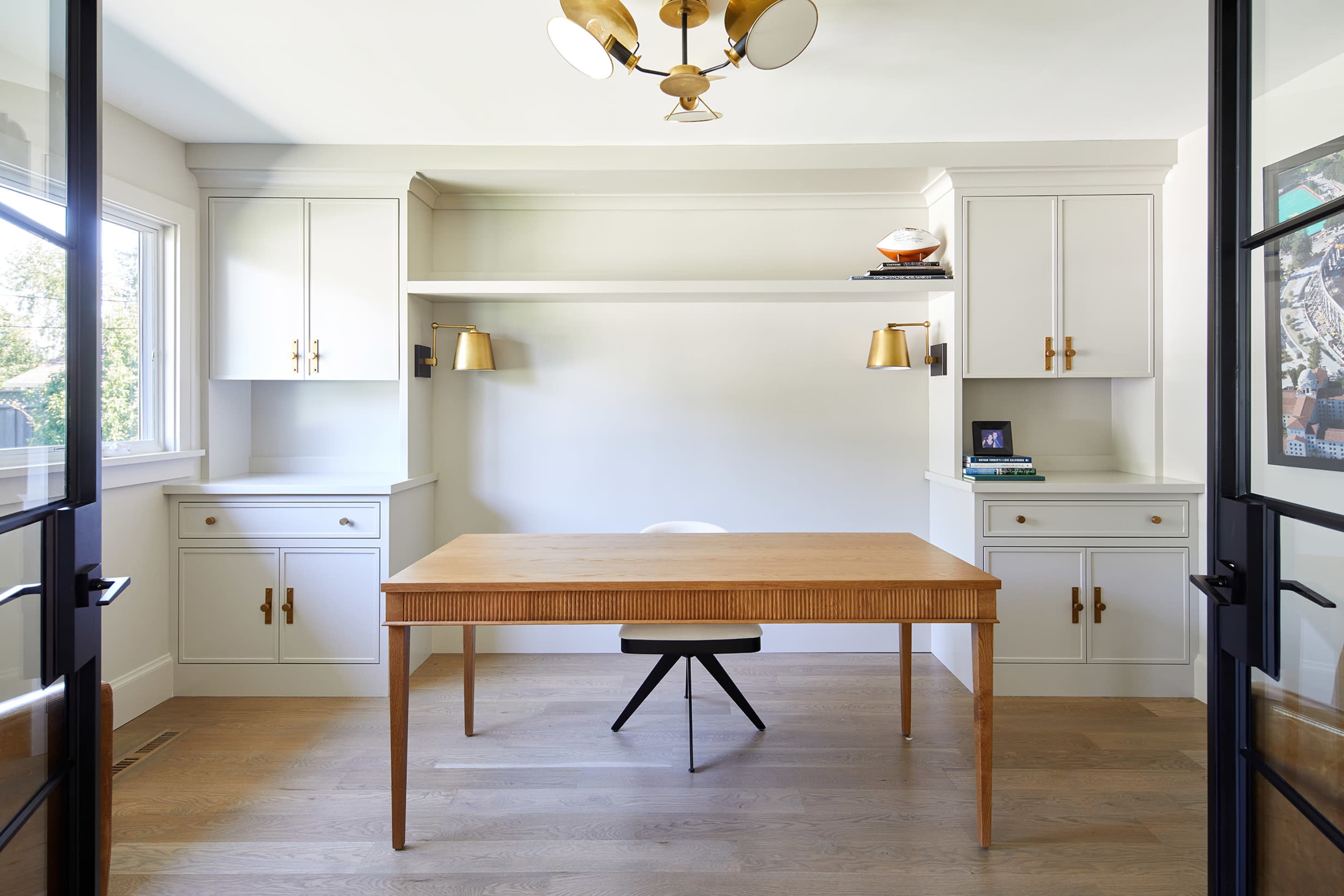 A modern home office features a central wooden desk with a black chair, flanked by built-in shelves and lamps, beneath a circular light fixture.