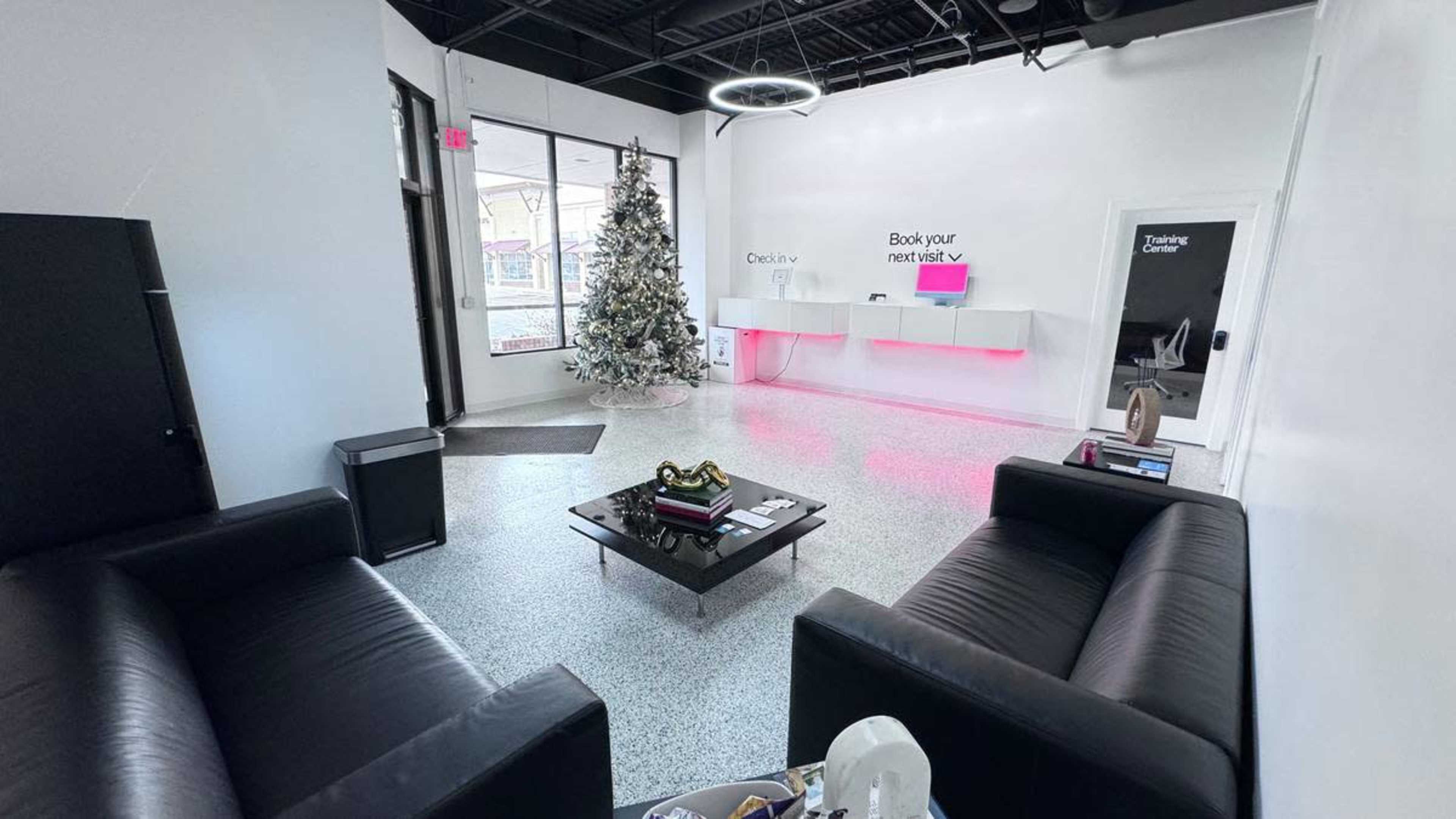 A modern reception area with two black sofas, a coffee table, a Christmas tree, and a white desk with pink lighting.