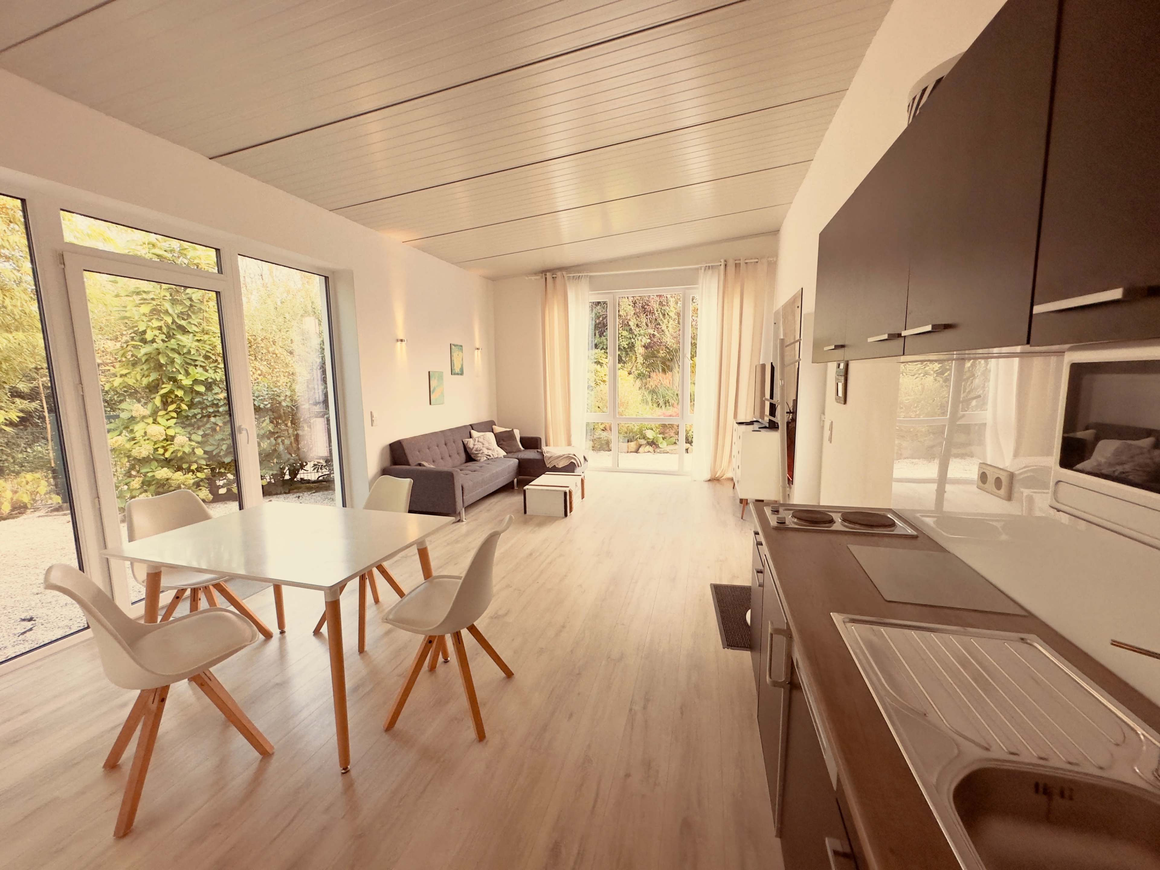 The image shows a modern, open-concept living space with a kitchen, dining area, and seating area, featuring large windows that let in natural light.