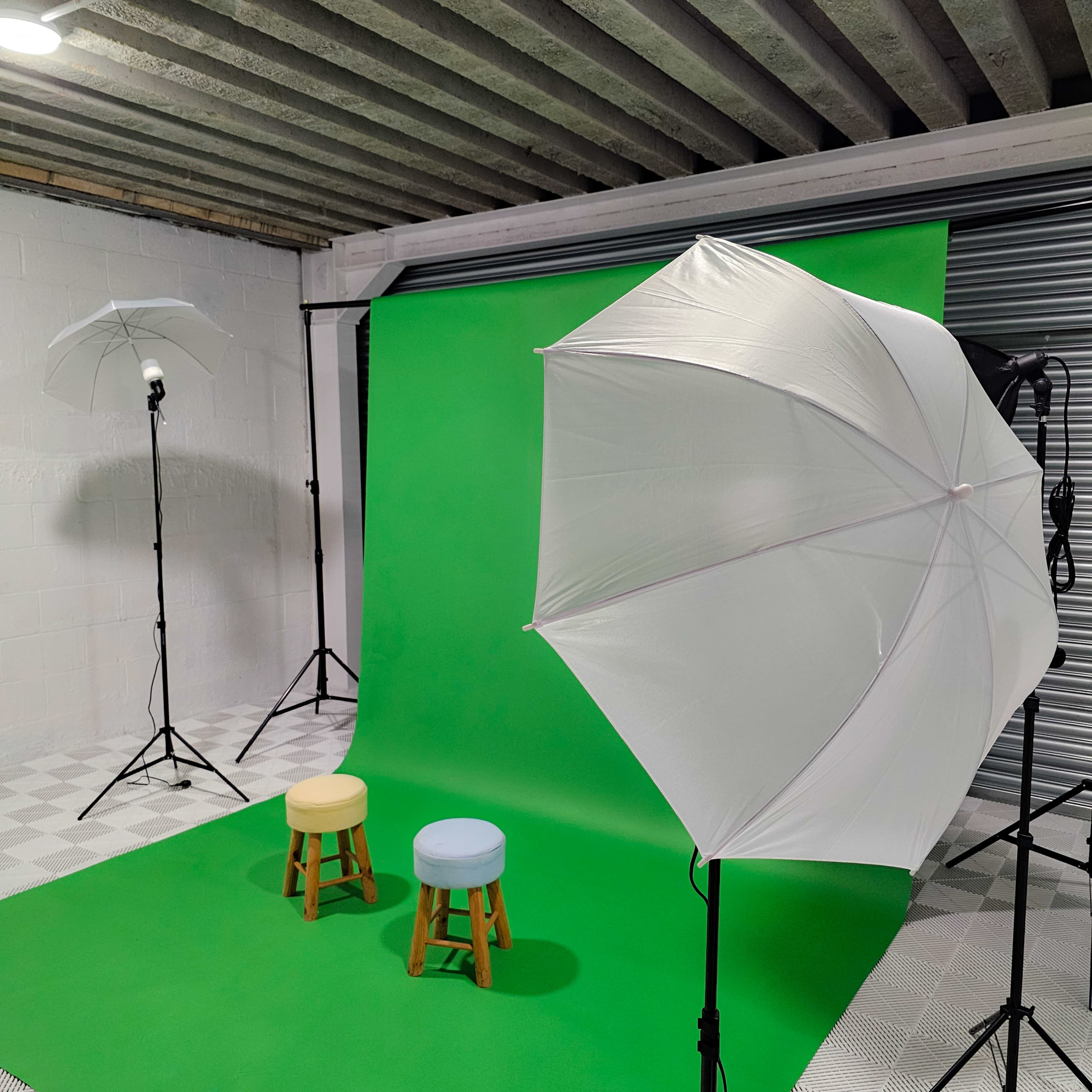 A photography studio with a green backdrop, two stools in pastel colors, and lighting equipment set up on either side.