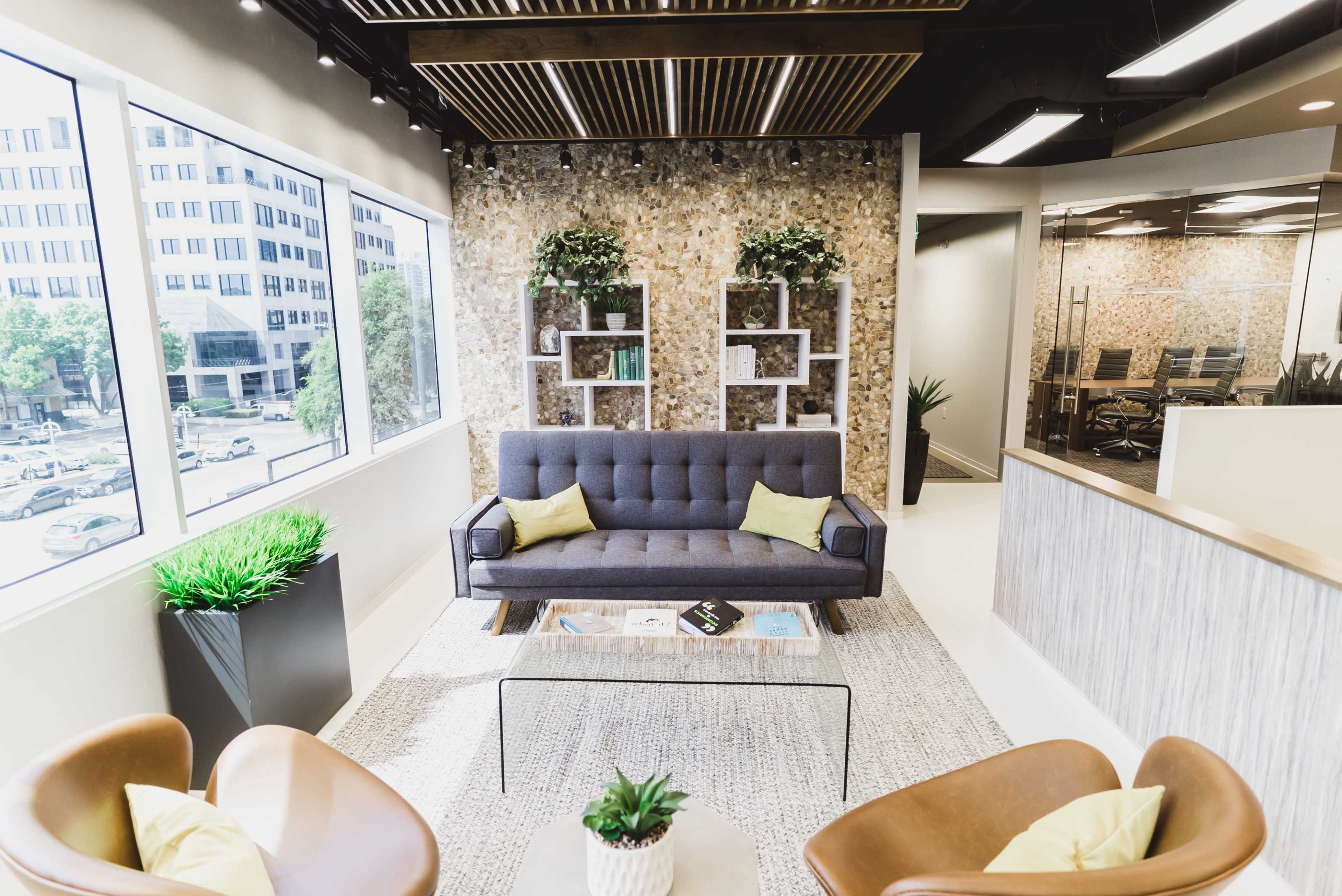 The image shows a modern office lounge area featuring a gray sofa with yellow accent pillows, two brown armchairs, a glass coffee table, and a decorative wall with plants and shelving.