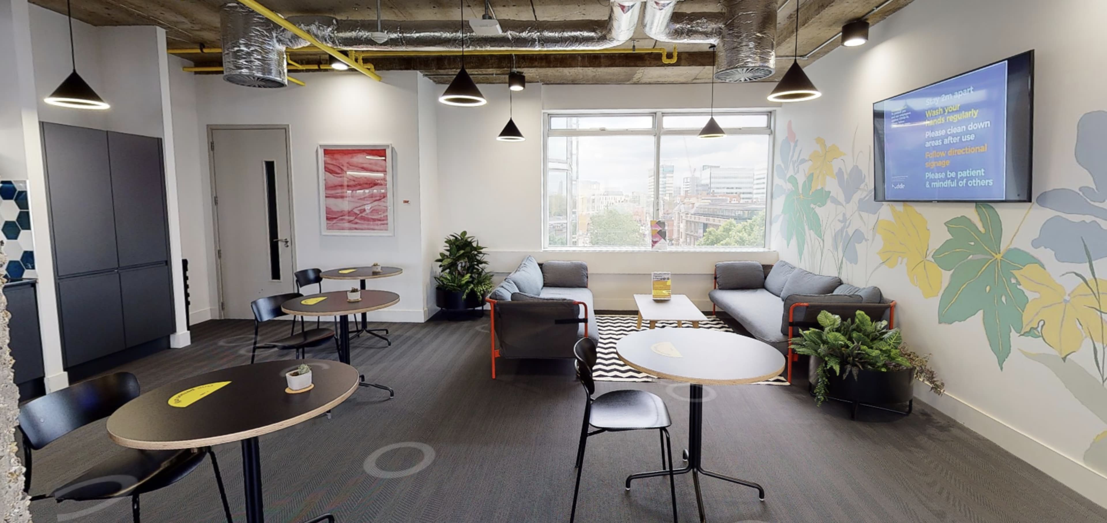A modern, bright office lounge area featuring several small tables, sofas, plants, and large windows with a cityscape view.